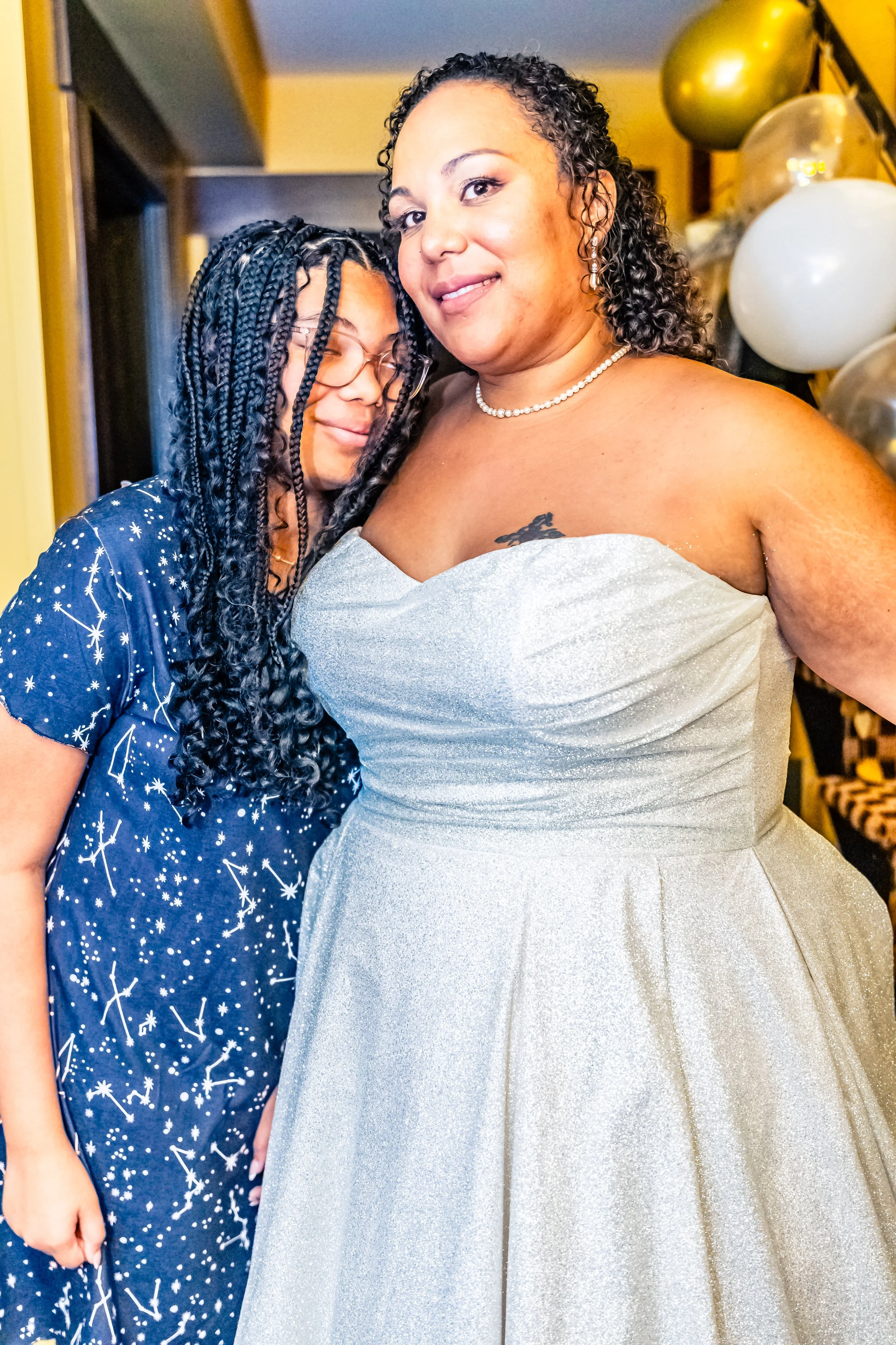 Two women hugging, one in a silver dress and the other in a navy star-patterned dress, at a celebration with balloons.