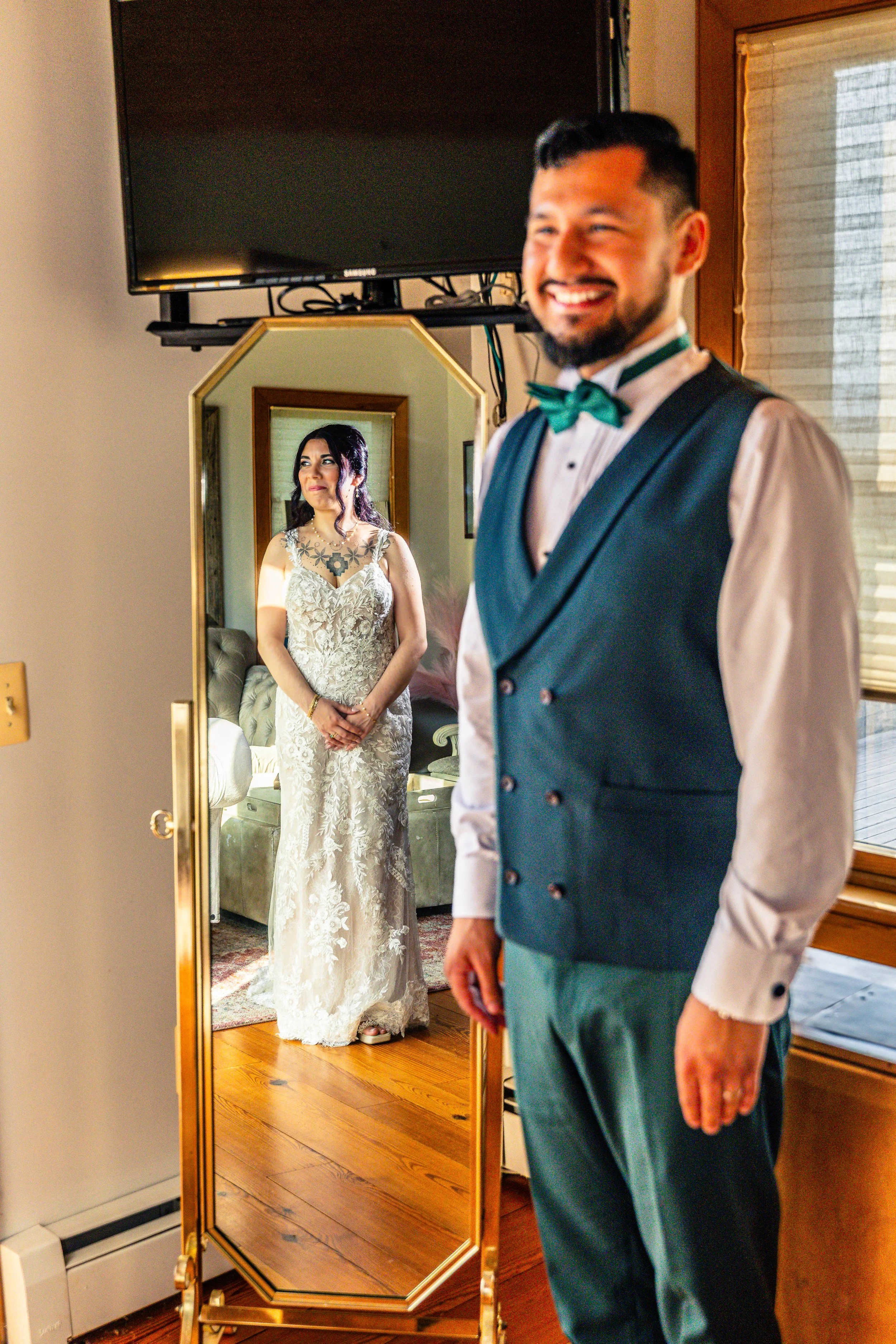 A groom looks at his bride through a mirror, with the bride standing behind him. The groom is smiling, dressed in a tuxedo with a green bow tie, and the bride is wearing a white lace wedding dress with tattoos visible on her chest and arms.