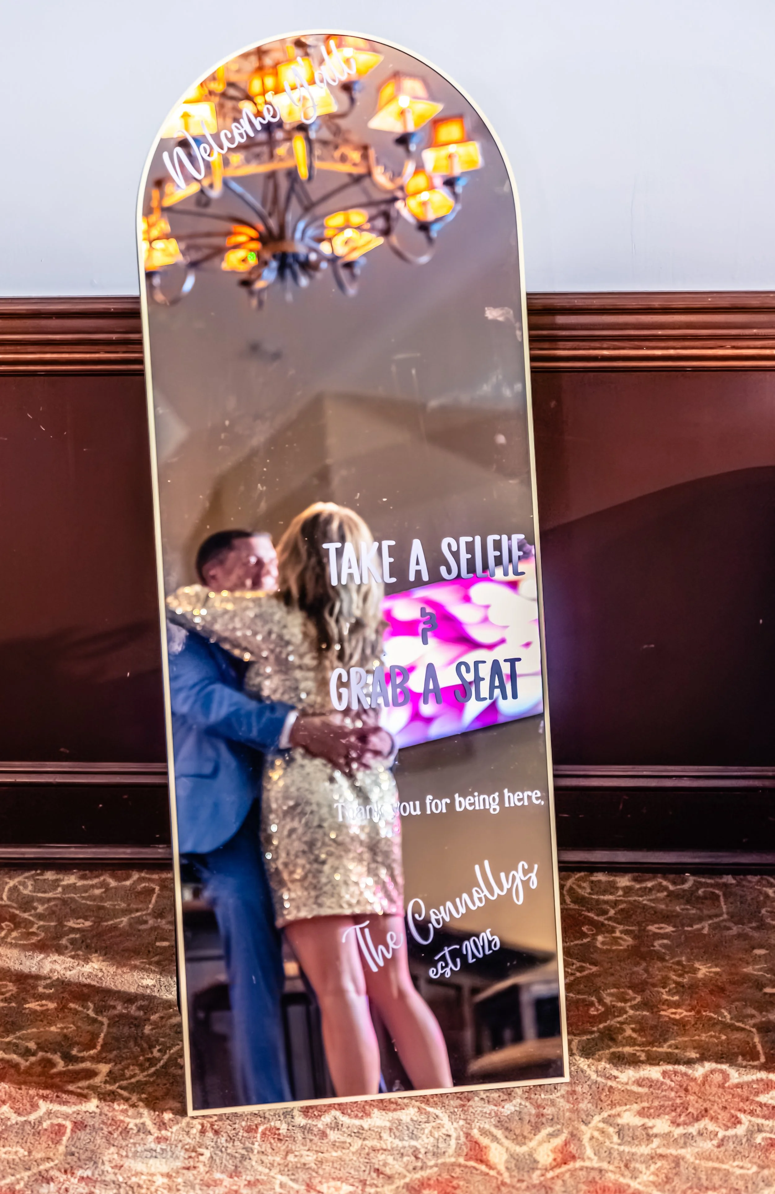 A decorative mirror with a reflection of a couple hugging, a chandelier above, and text that reads "Welcome to the party, take a selfie, grab a seat."