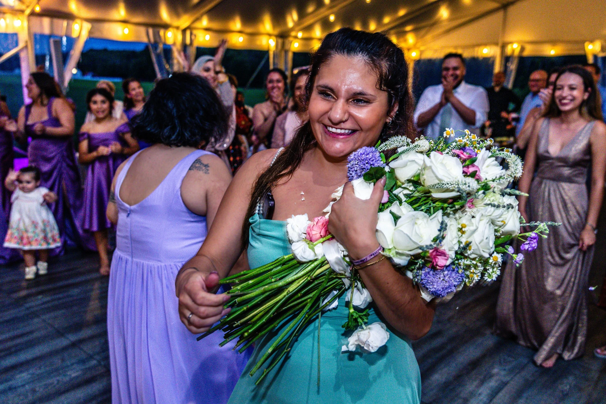 A woman in a teal dress holding a large bouquet of white, pink, purple, and green flowers, smiling at a celebration or party