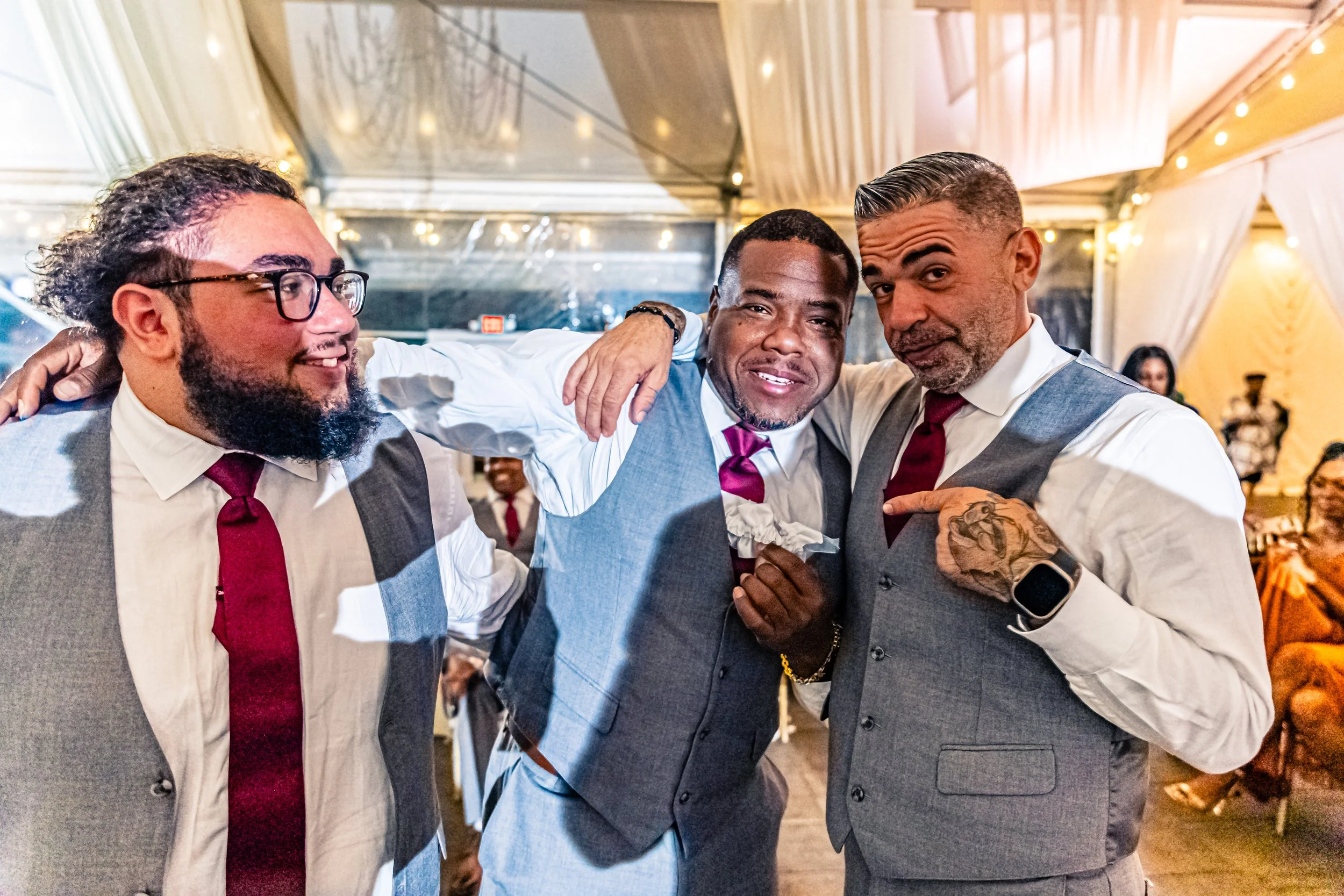 Three men in suits with ties, smiling and posing together at an indoor event, with other guests seated in the background.