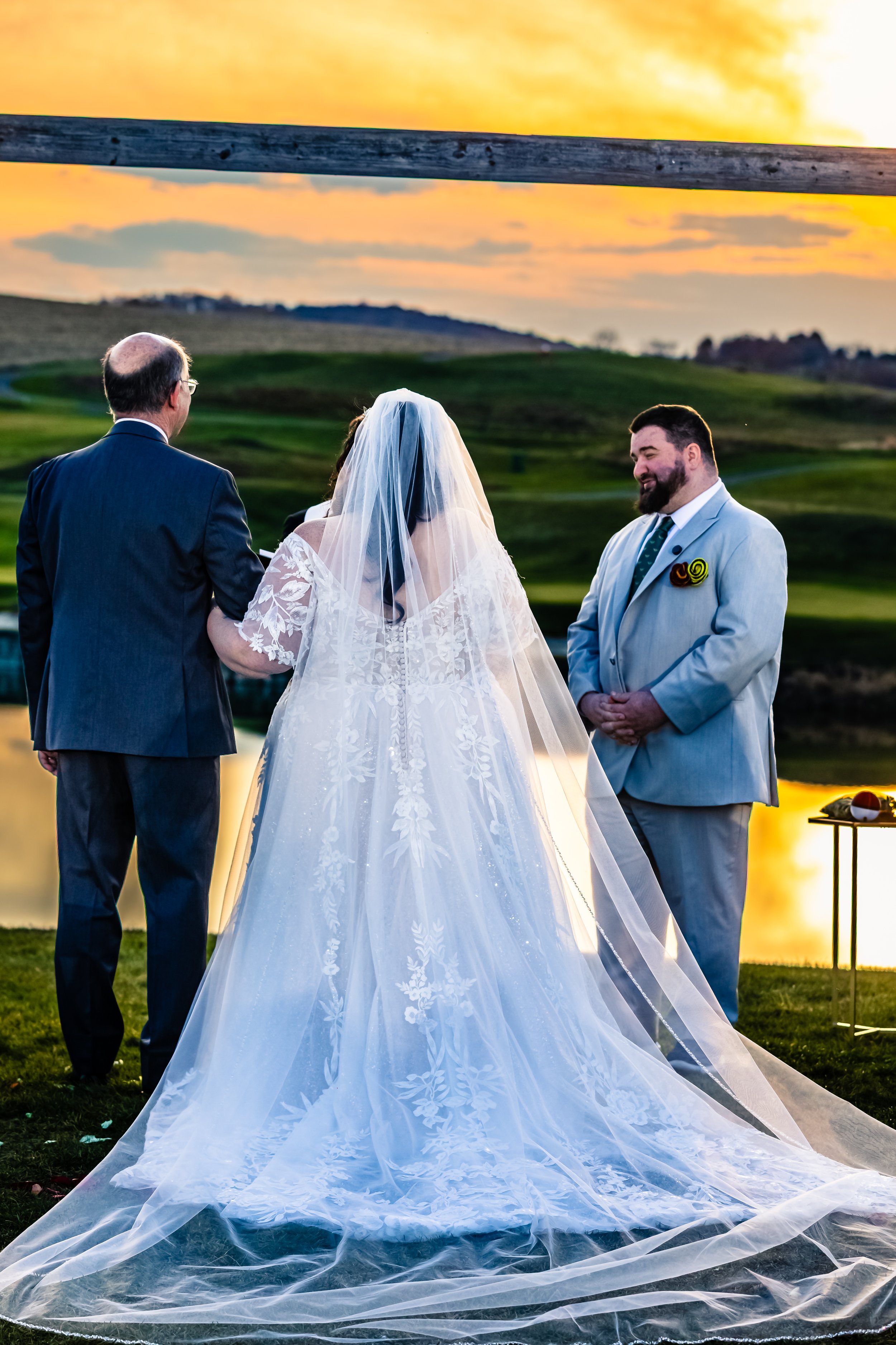 A wedding ceremony taking place outdoors during sunset, with a bride in a white gown and veil, a groom in a light gray suit, and an officiant, by a lake with a scenic landscape in the background.