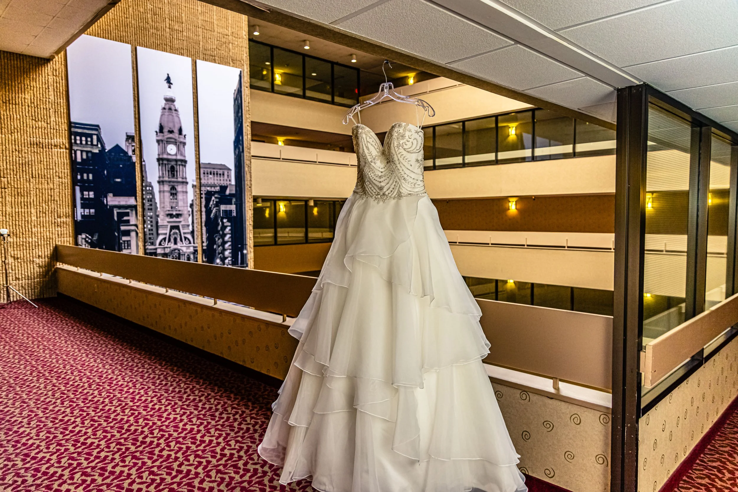 Bridal wedding dress hanging on a clear hanger inside a hotel or convention center.