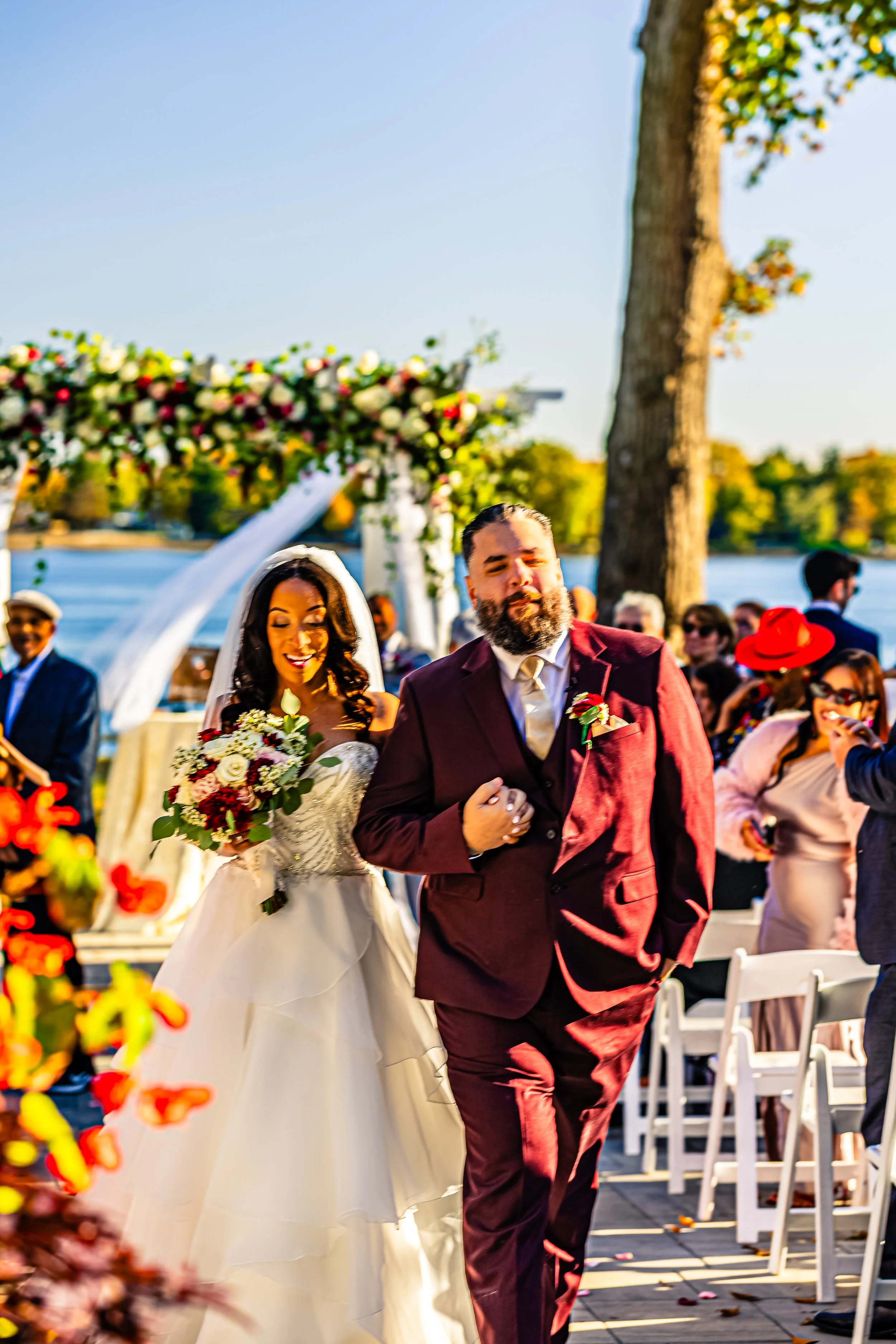 A bride holding a bouquet walking down the aisle with a groom during an outdoor wedding ceremony by a lake, surrounded by guests and decorated with floral arrangements.