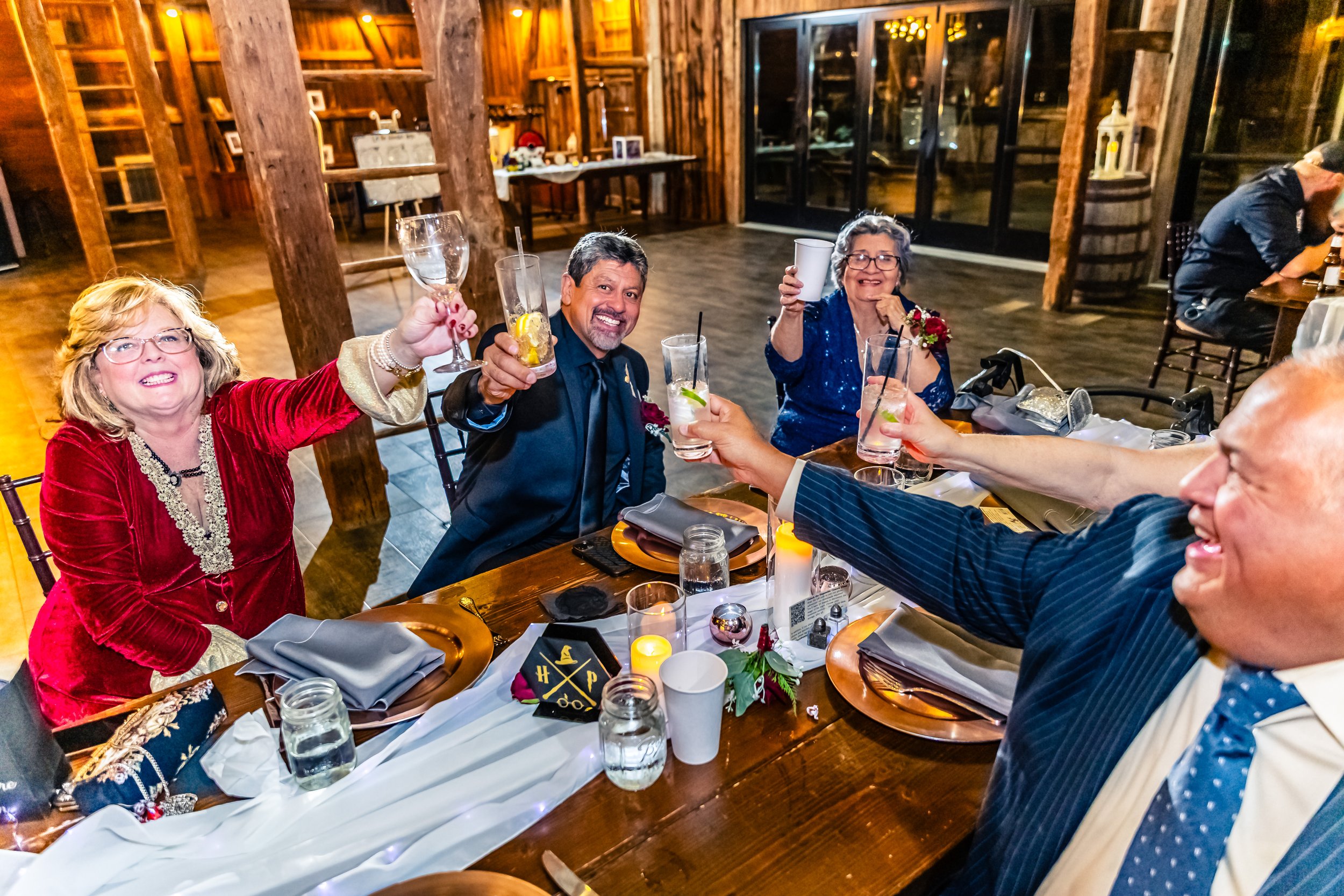 Group of five adults celebrating at a dinner table, raising glasses in a toast, smiling and enjoying drinks, in a warmly lit rustic venue with wooden walls and large windows.