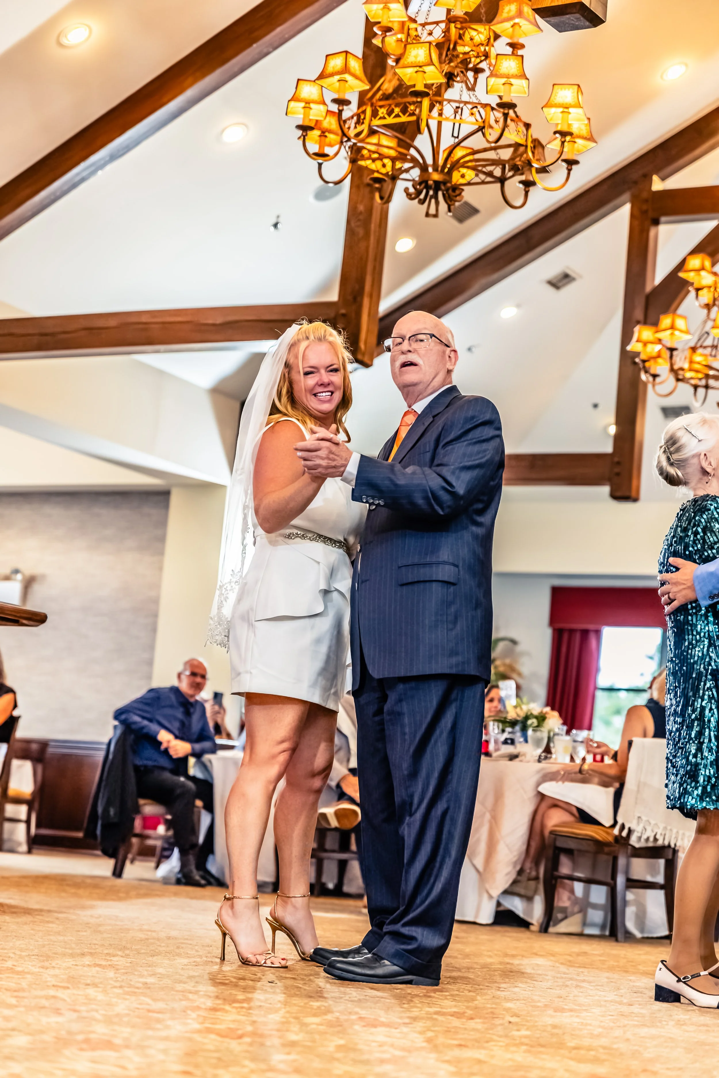 A woman in a white dress and high heels dancing with an older man in a dark suit at a wedding reception, with onlookers seated at tables in the background.