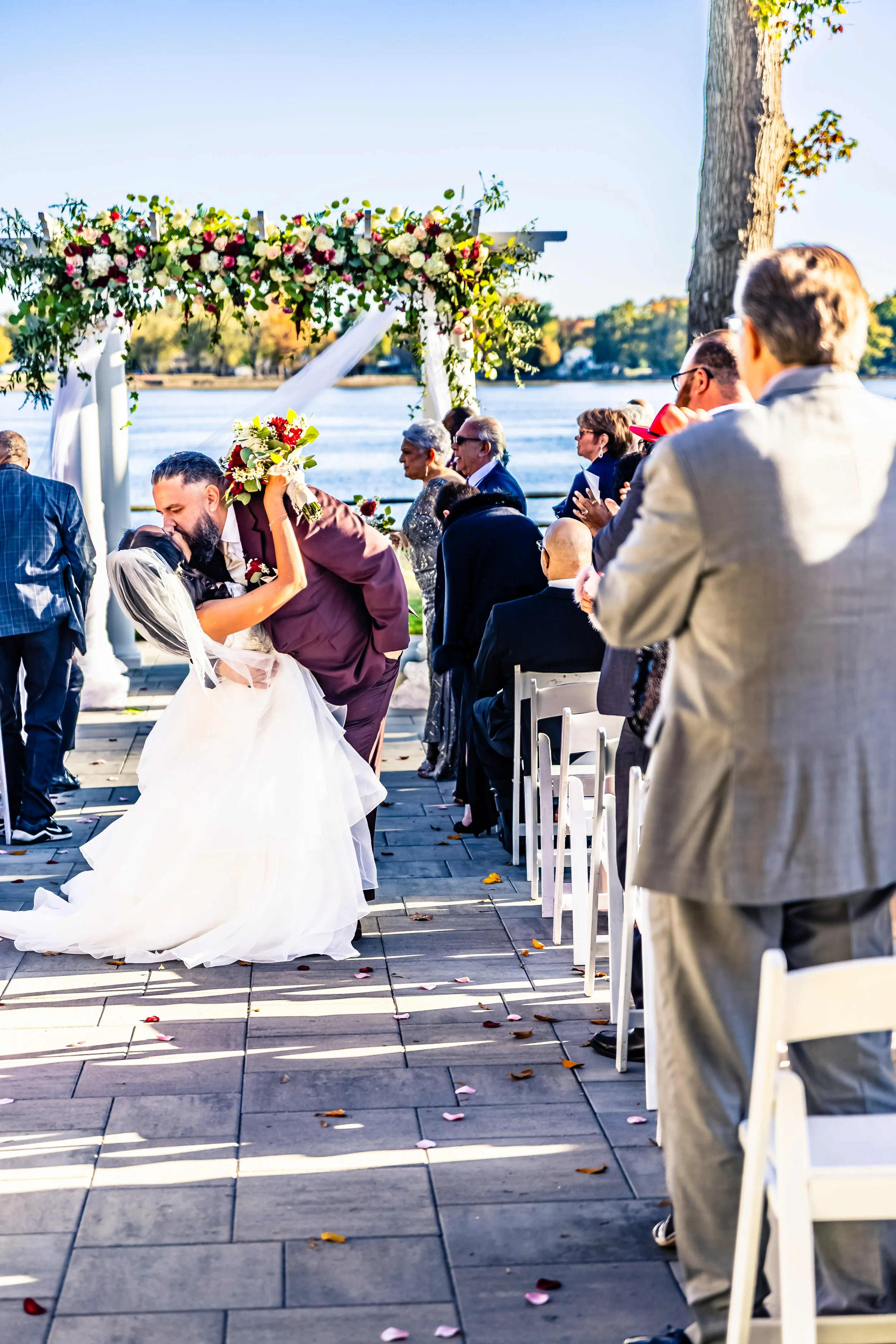 A wedding ceremony outdoors by the water, with the bride and groom sharing a kiss under a floral arch, surrounded by seated guests.