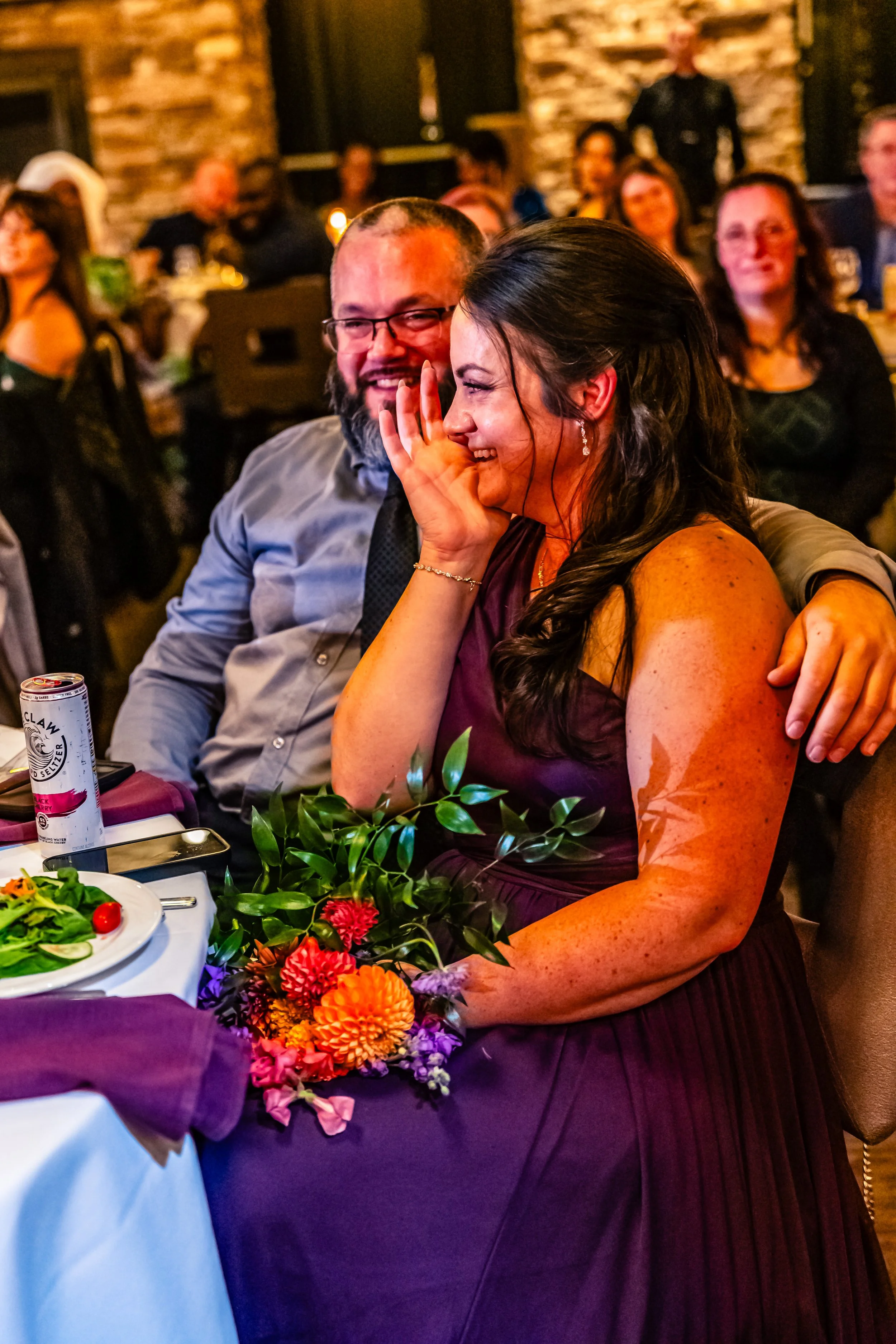 A woman in a purple dress and a man in a gray shirt are sitting closely, laughing and sharing a joyful moment at a celebration or wedding reception. The woman holds a bouquet and covers her mouth while laughing. Other guests are visible in the backgr