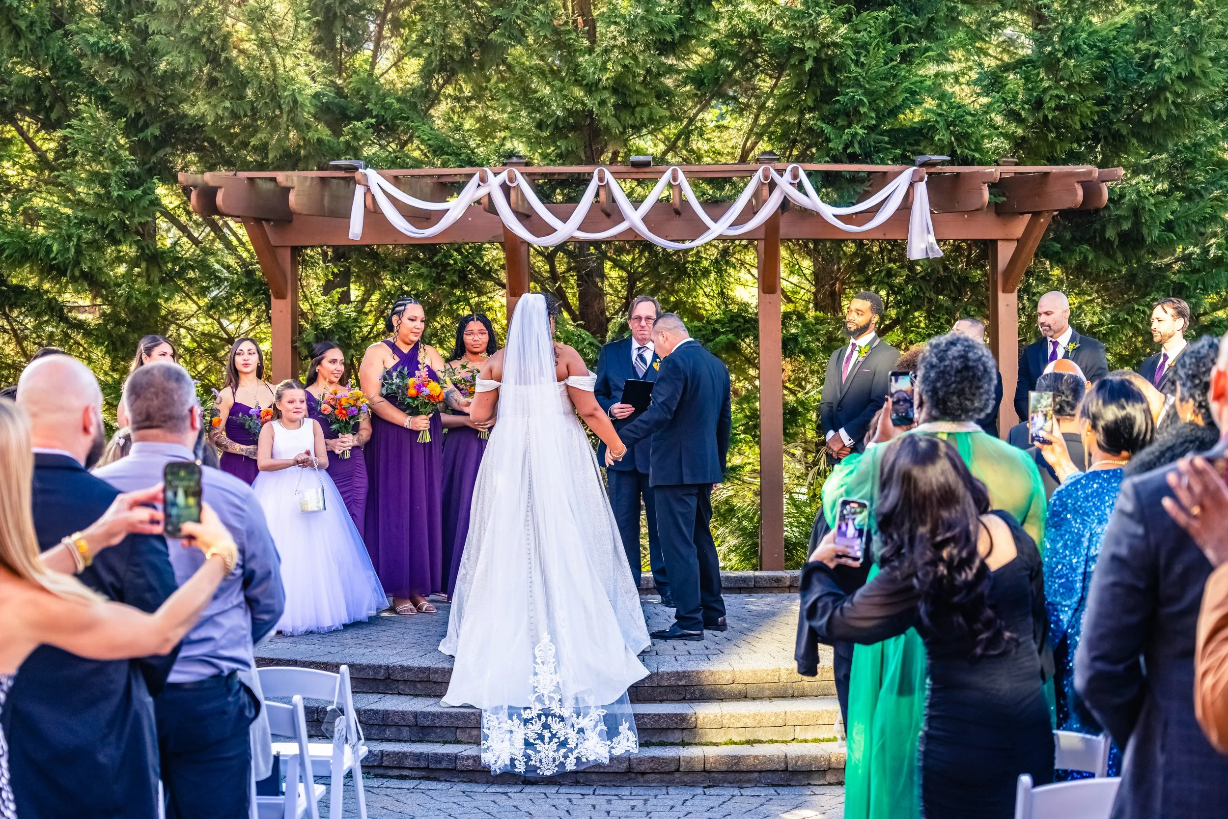 A wedding ceremony outdoors on a small stage, with a bride and groom exchanging vows, surrounded by officiant and wedding party, with guests taking photos in front of lush green trees.