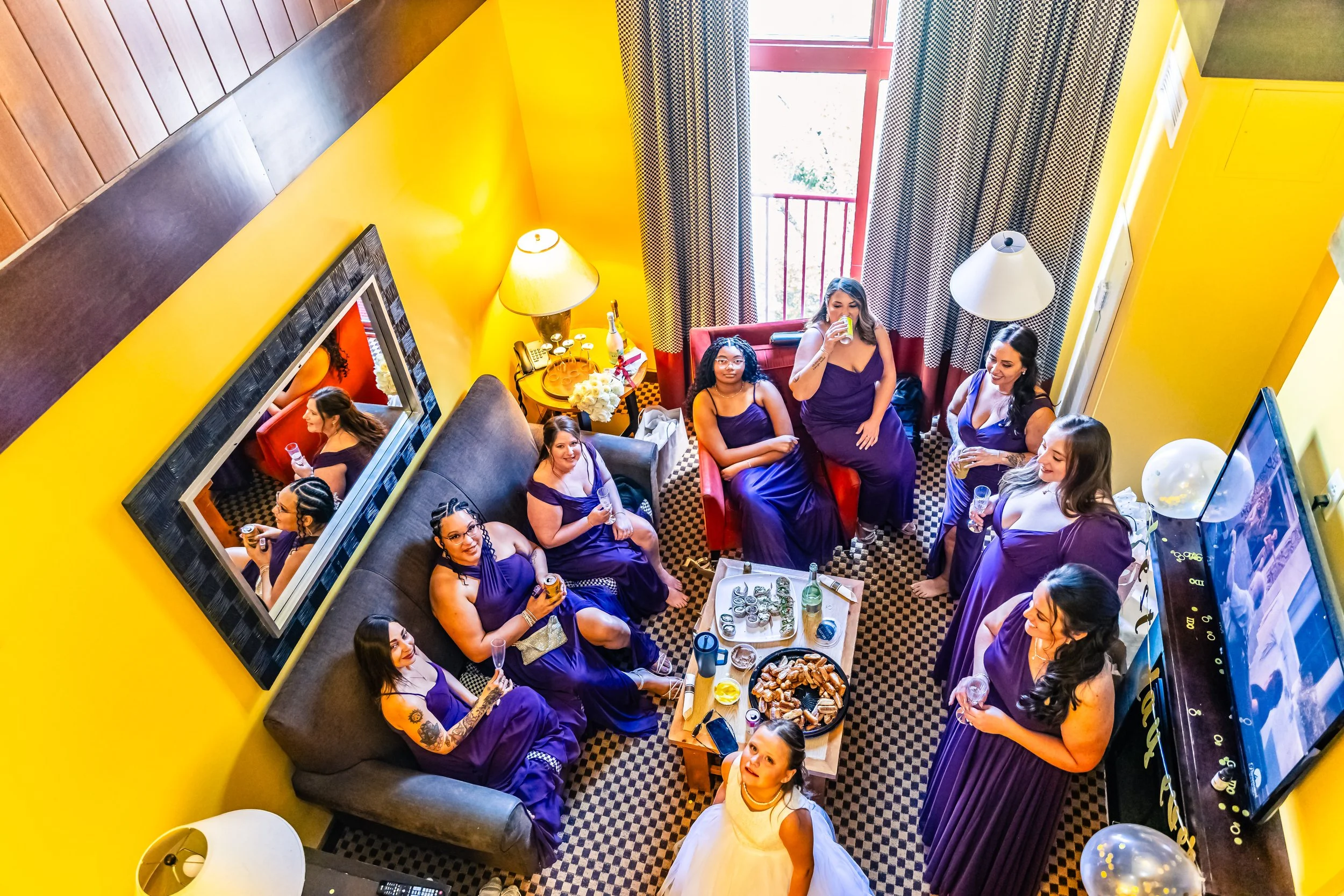 A group of women in purple bridesmaid dresses gathered in a colorful living room, enjoying drinks and snacks during a wedding preparation, with a bride in a white dress among them.