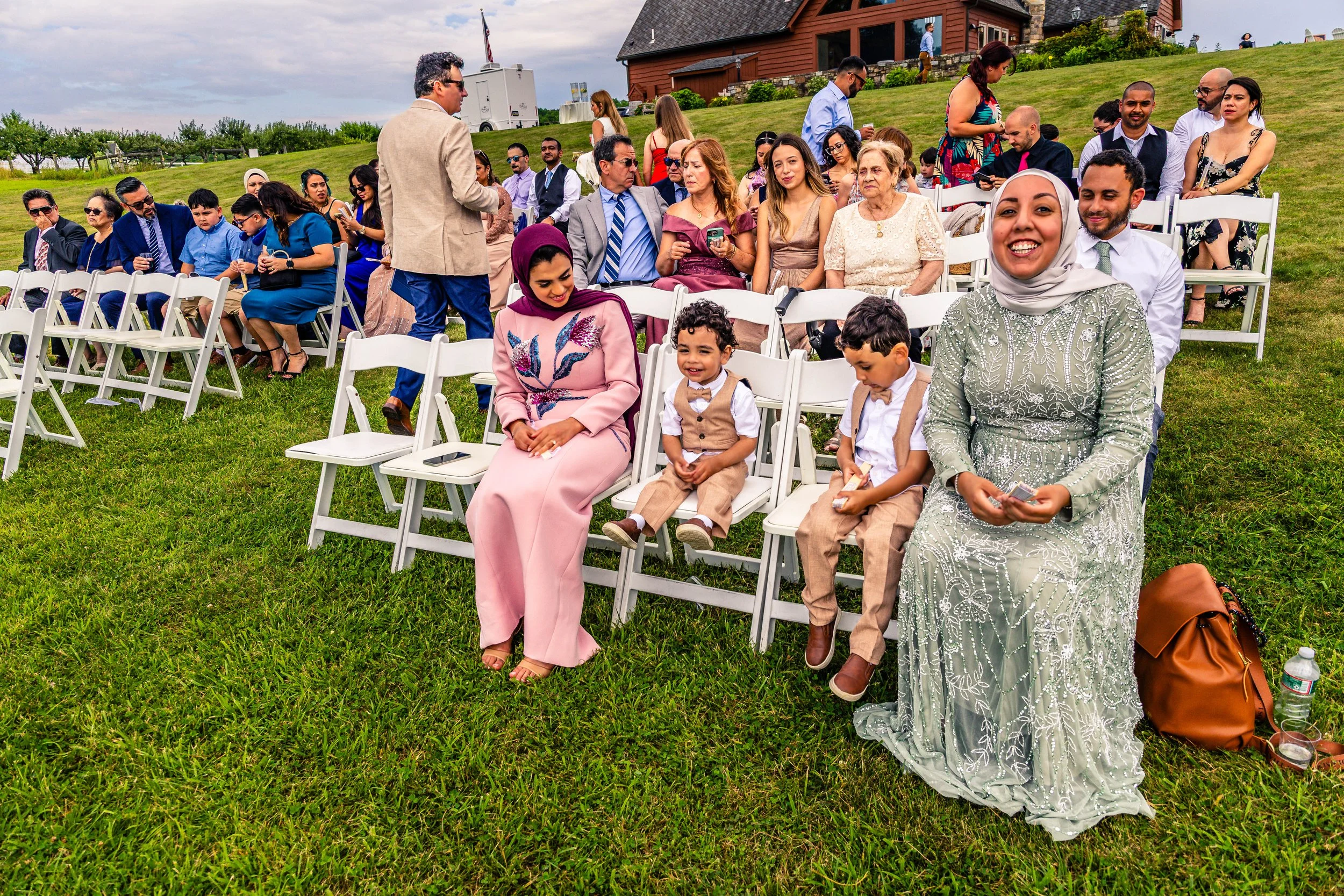 Group of people sitting on white folding chairs outdoors on a grassy area, attending a celebration or event with a building and trees in the background.