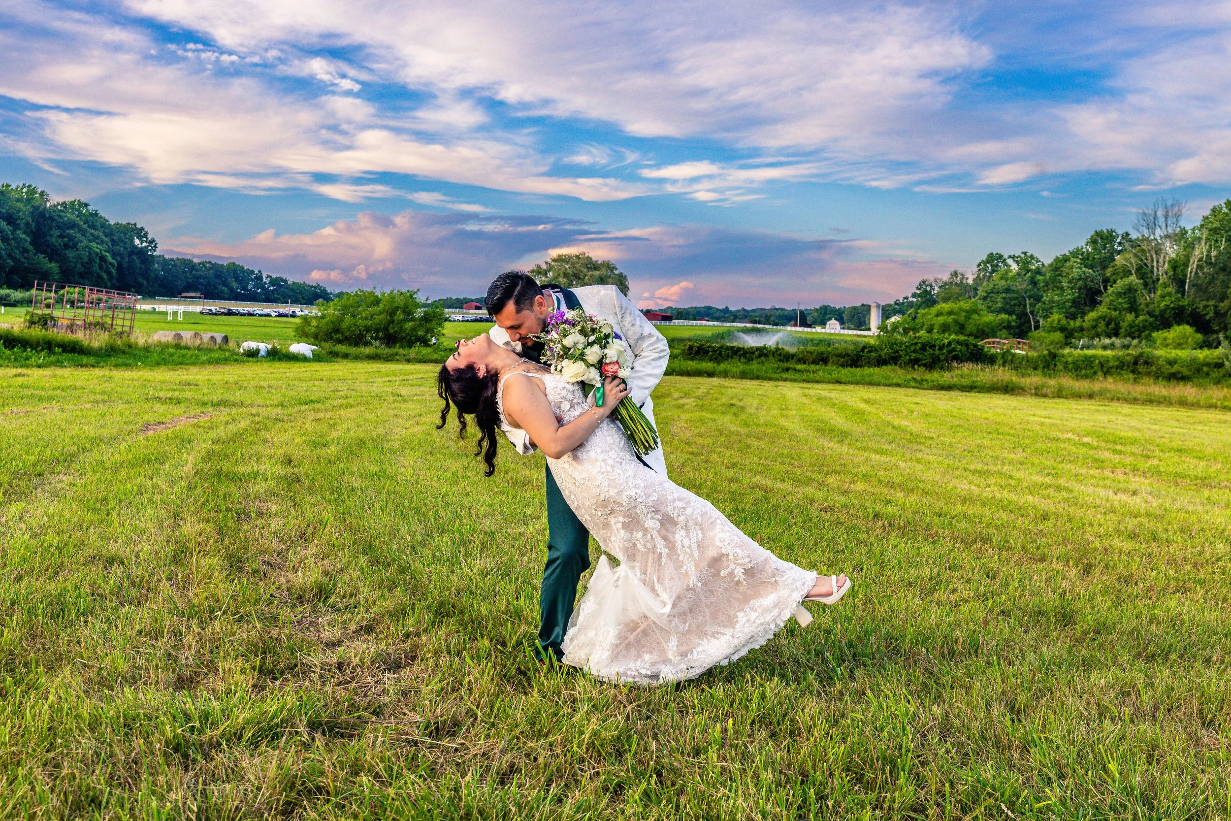 A bride and groom in wedding attire dancing or celebrating outside on a green field at sunset with a scenic sky and farm in the background.