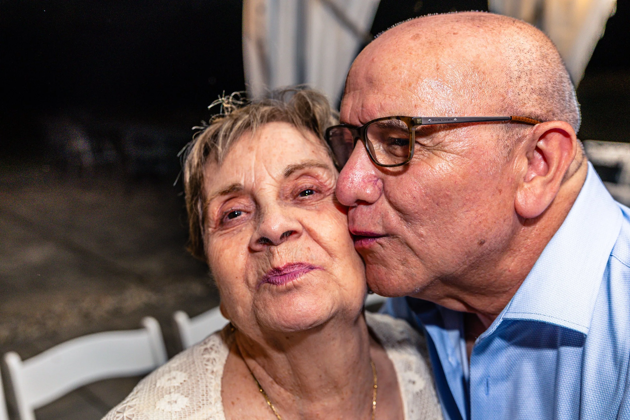 An elderly woman with short light brown hair and a necklace receiving a kiss on the cheek from a man with glasses and a bald head, both smiling softly.