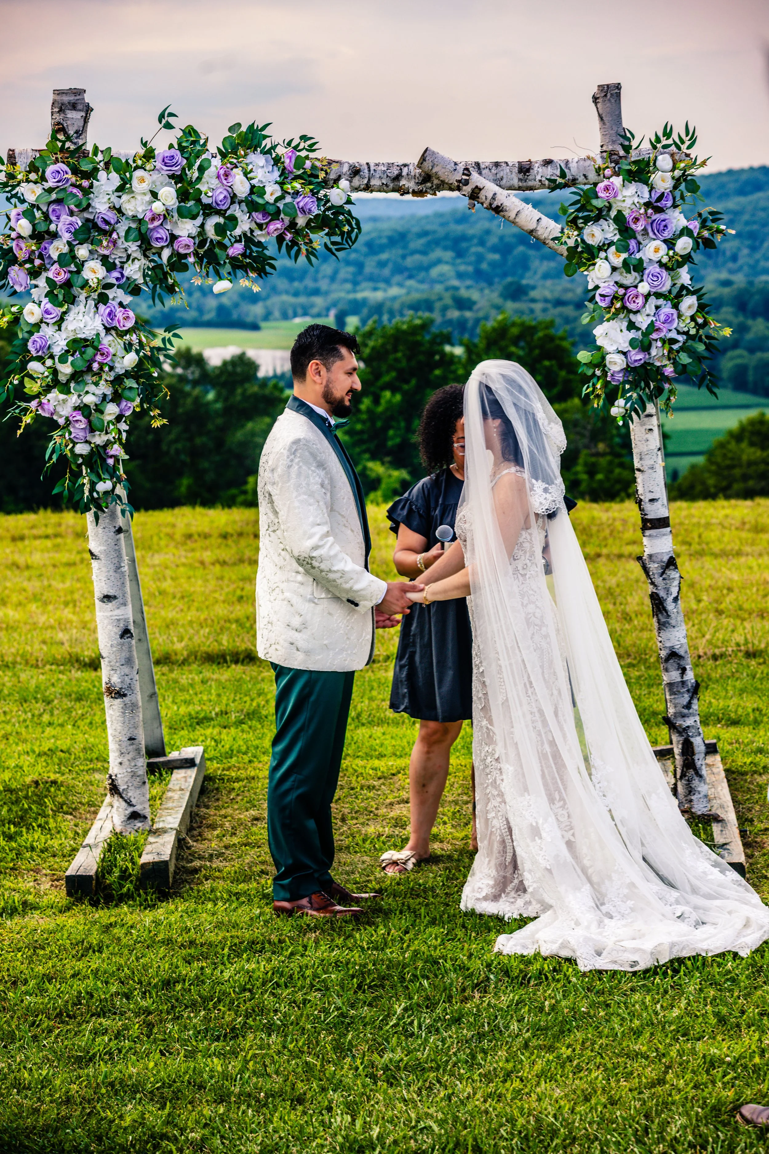 A couple getting married outdoors under a flower-adorned wedding arch in a green field, with a scenic landscape and hills in the background.