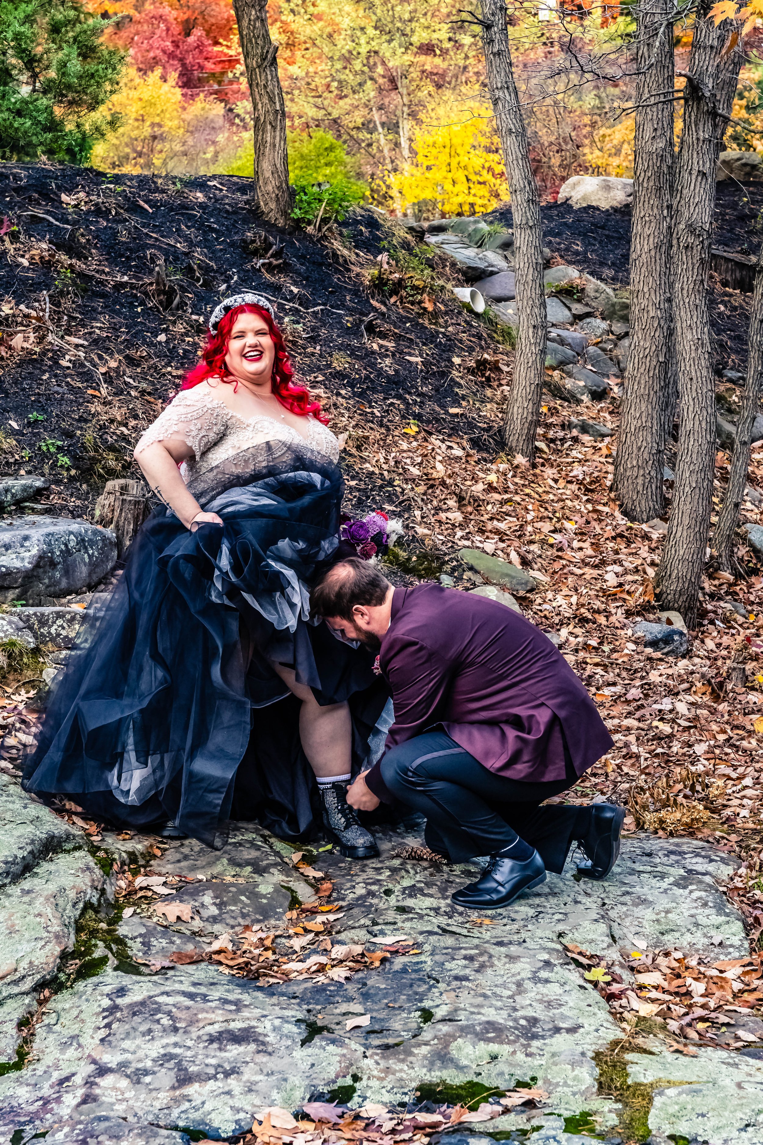 A woman in a formal gown with red hair and a crown, standing outdoors on rocks with trees and colorful fall foliage in the background, while a man kneels, helping her with her shoe.