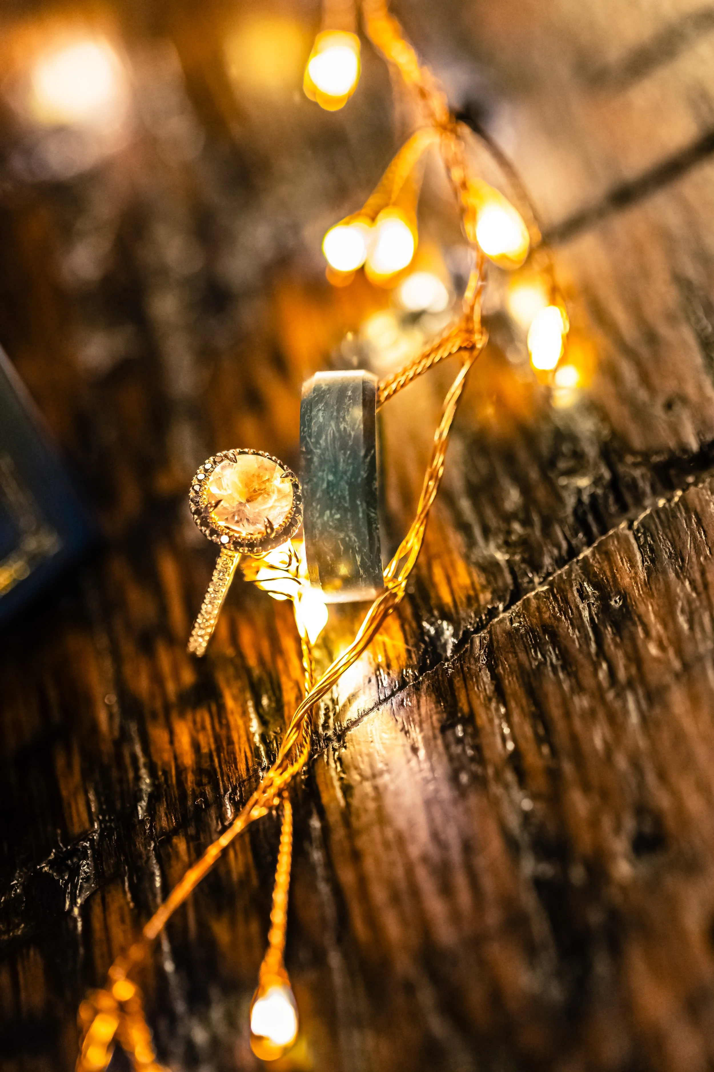 Close-up of a gold ring with a large yellow gemstone and smaller surrounding stones, hung on a string of warm fairy lights on a wooden surface.