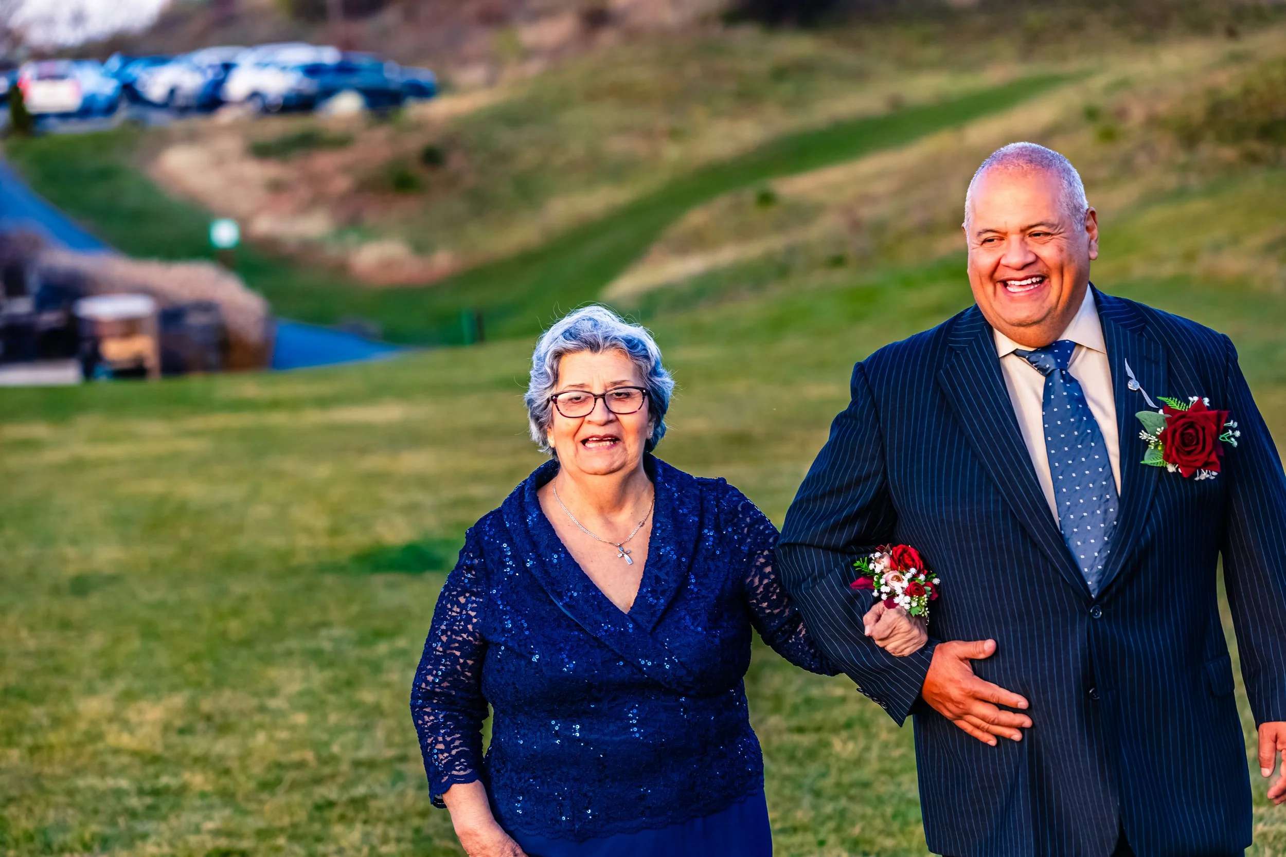 An elderly woman in a blue lace dress and glasses, and an elderly man in a navy pinstripe suit with a blue polka dot tie, smiling and holding hands outdoors on a grassy hill with a parking lot and playground in the background.