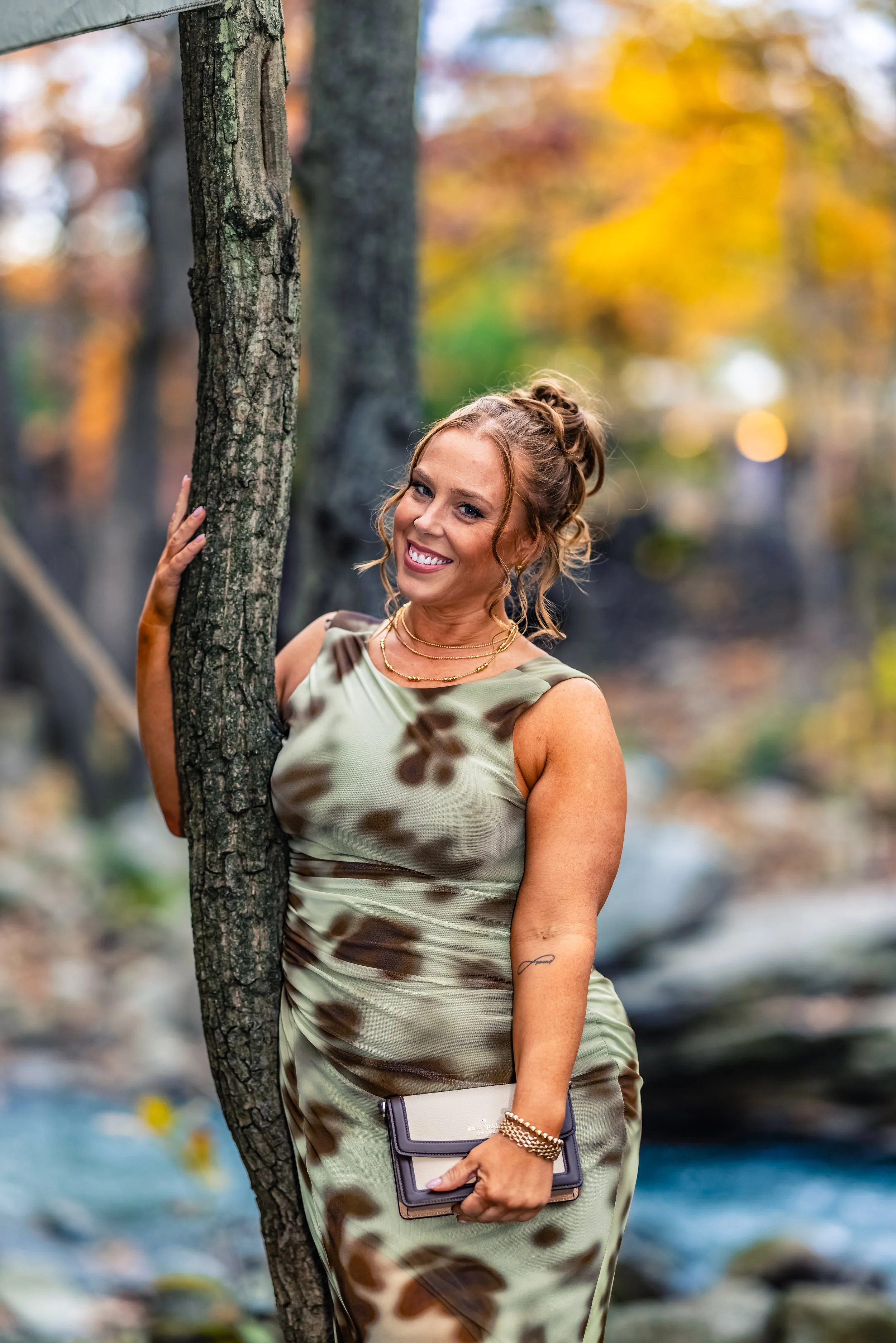 A woman with curly red hair, wearing a sleeveless dress with a brown and green tie-dye pattern, standing outdoors next to a tree with a blurred background of autumn foliage and a water stream, smiling and holding a small clutch purse.