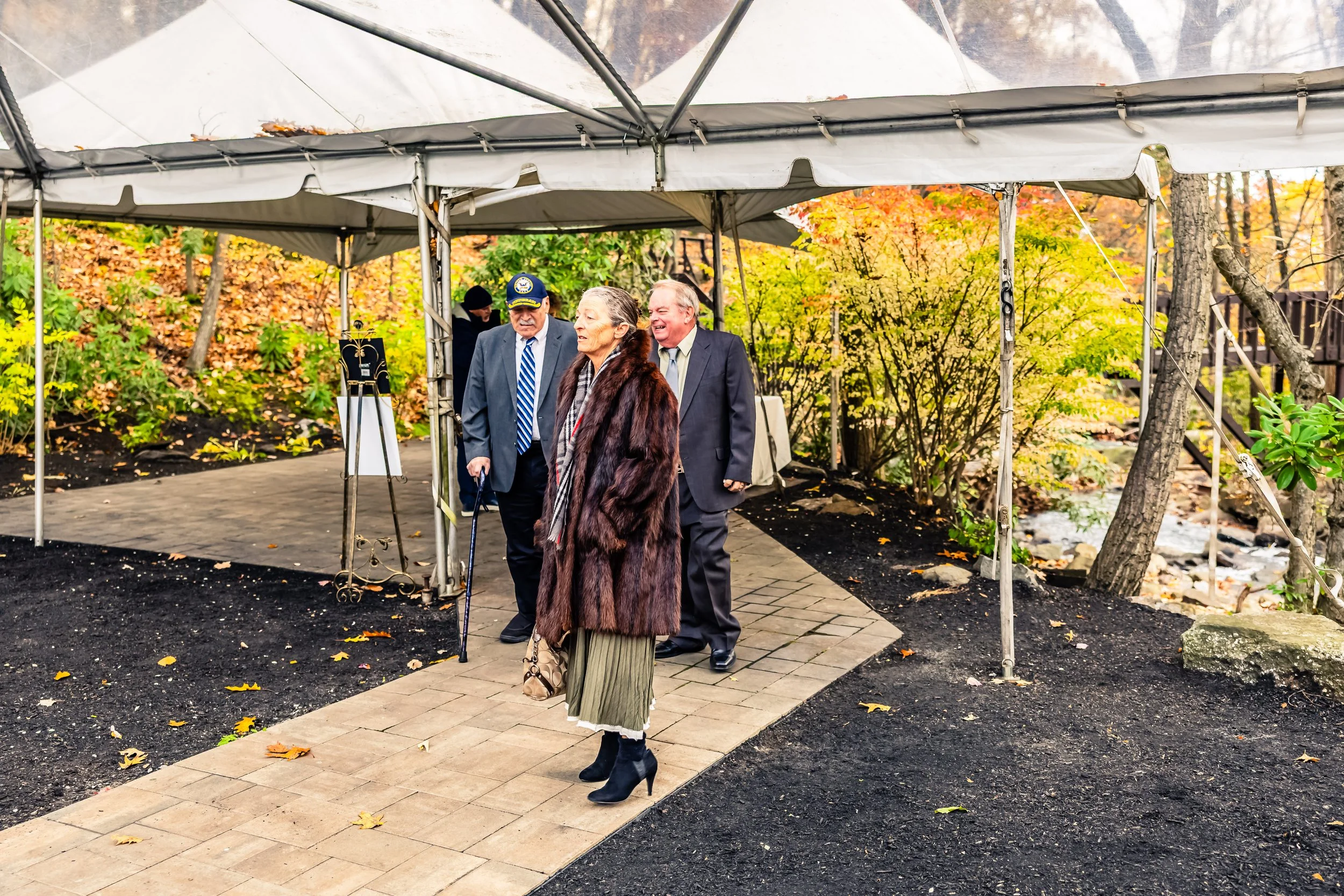 Three people walking under a canopy in a park-like area with autumn foliage, two men in suits and a woman in a fur coat, one man wearing a hat, and the woman carrying a handbag.