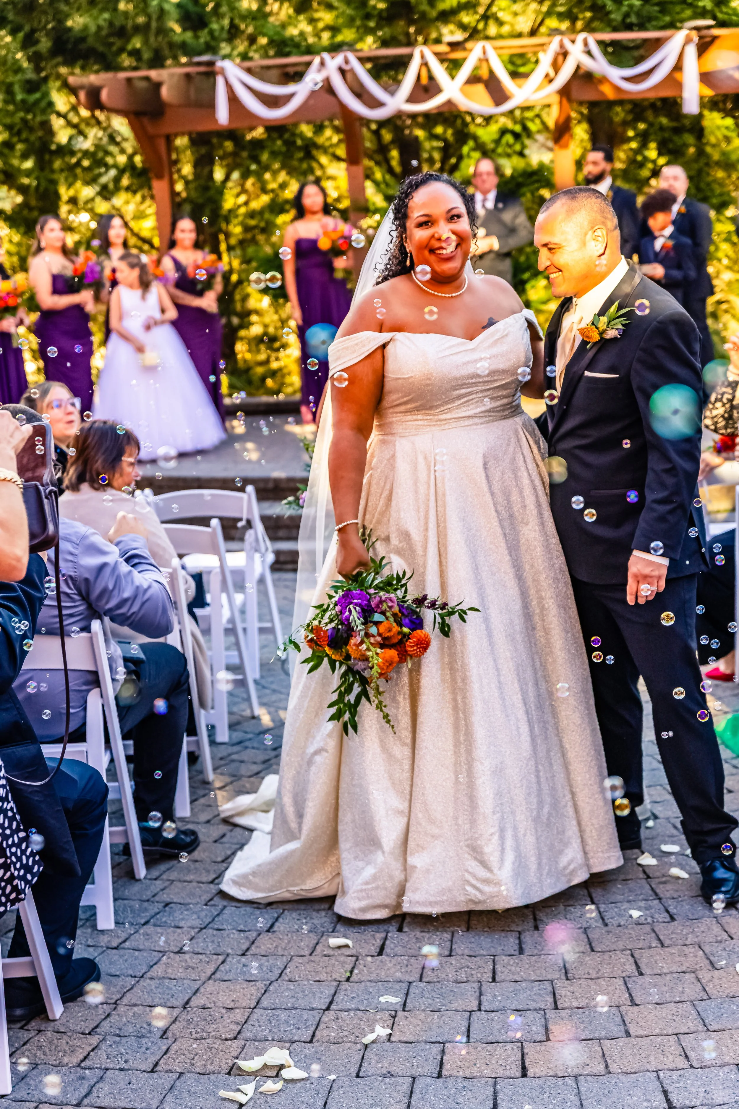 Bride and groom smiling at each other at an outdoor wedding ceremony, surrounded by guests and bridesmaids in purple dresses, with bubbles floating in the air.
