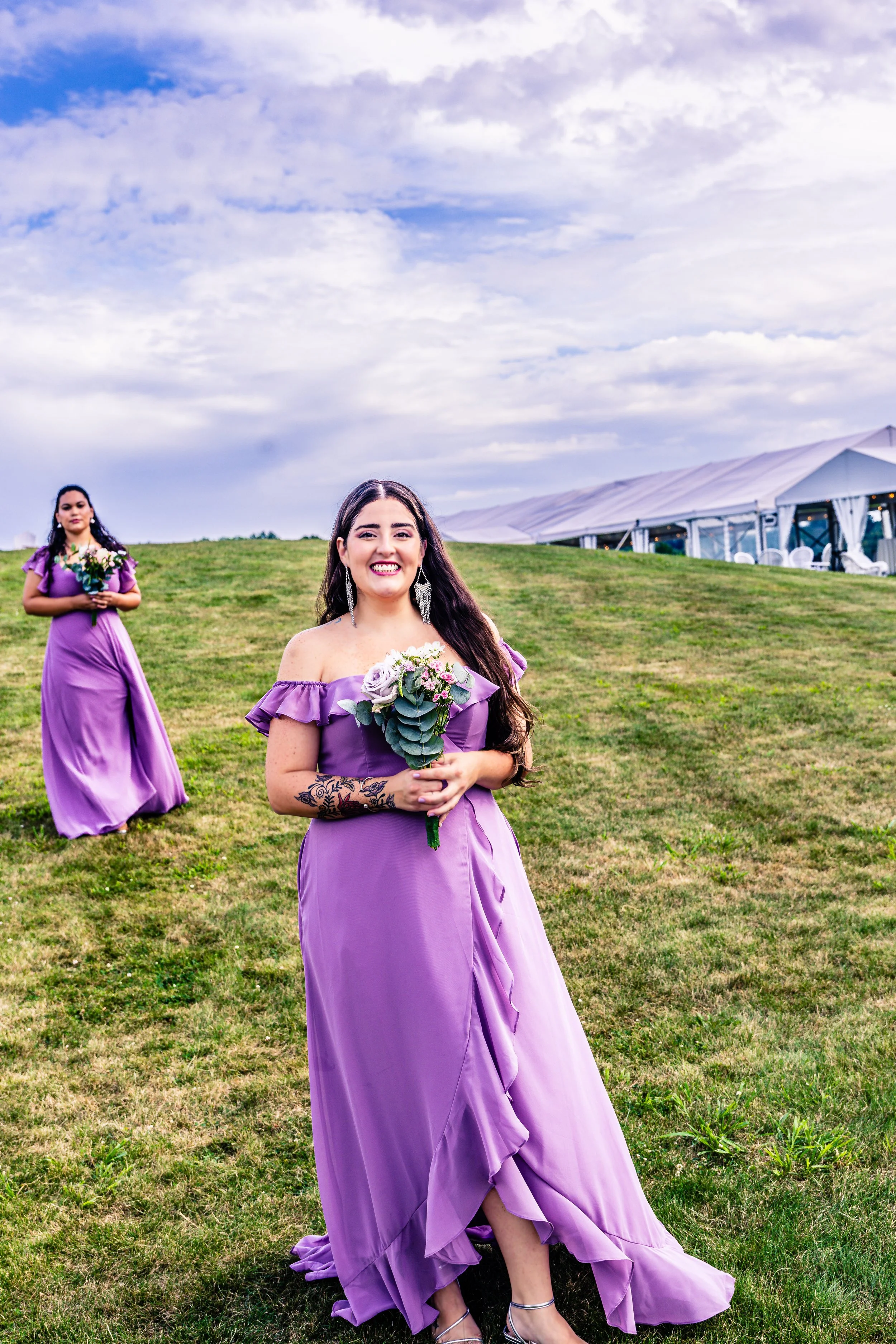 A woman in a purple dress smiling and holding a bouquet of flowers, standing on grass with another woman in a similar dress holding a bouquet in the background, under a cloudy sky with a large tent in the distance.