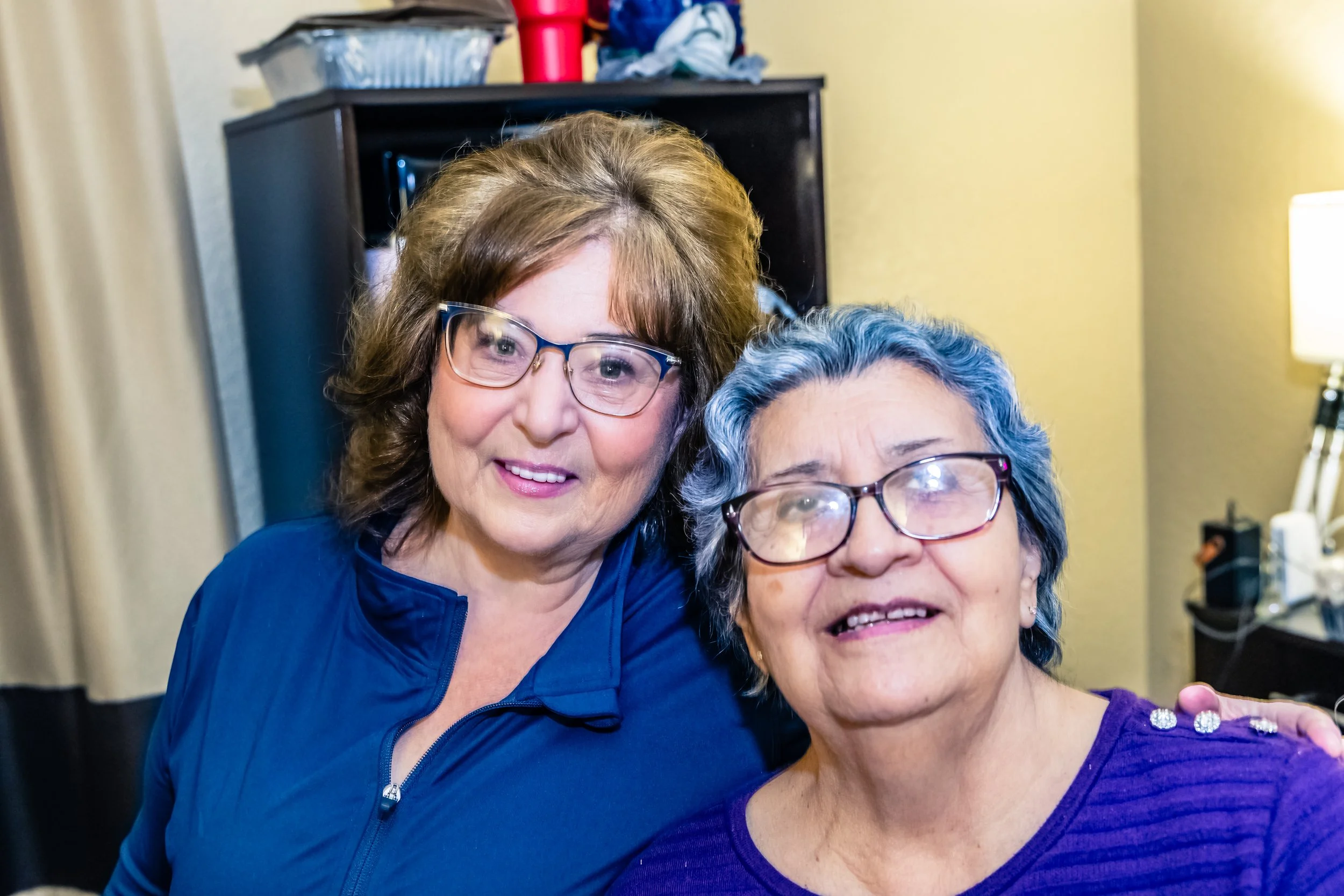 Two women with glasses smiling and posing together in a warmly lit room.