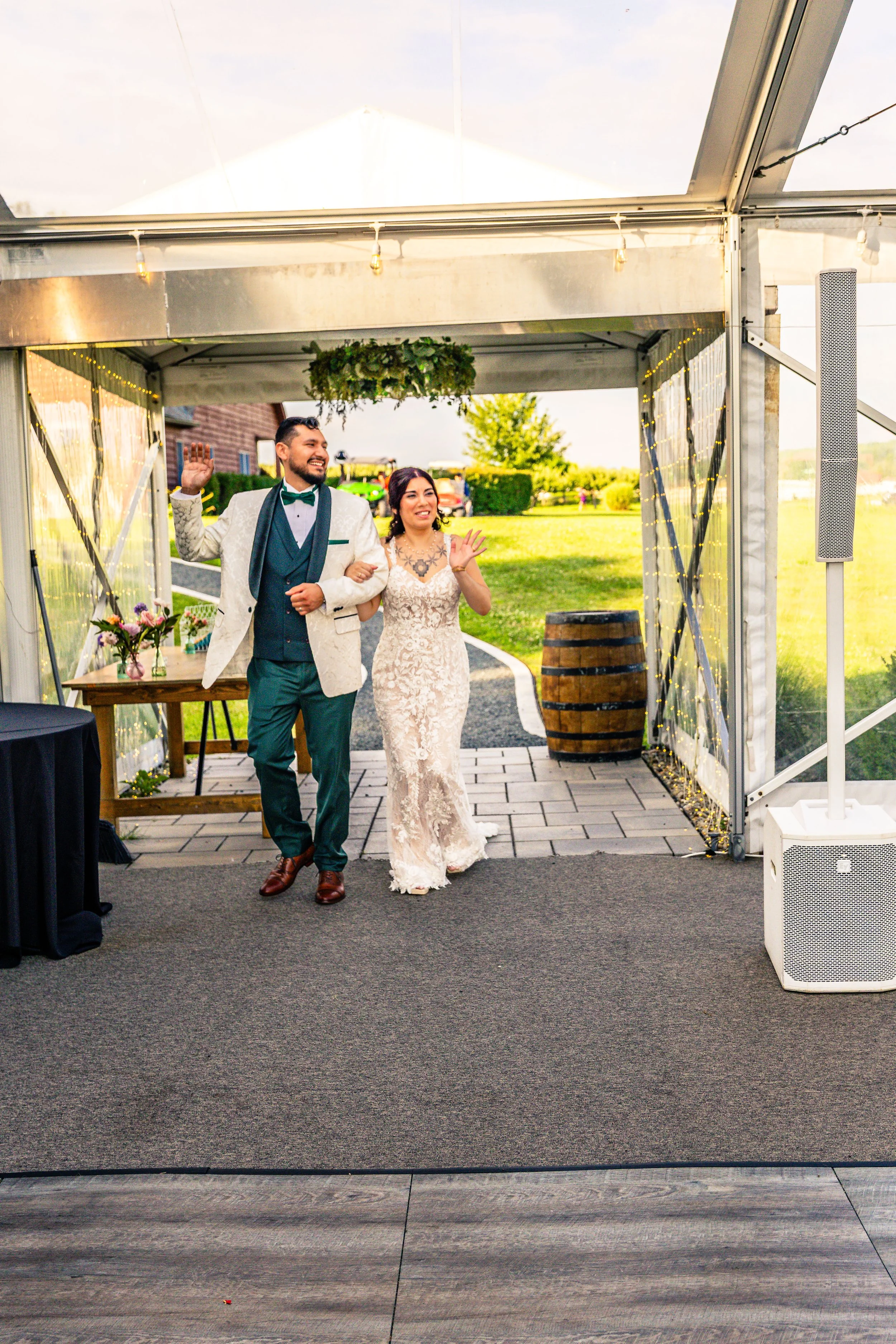 A bride and groom walking into a wedding reception tent, waving and smiling with lakeside scenery in the background.