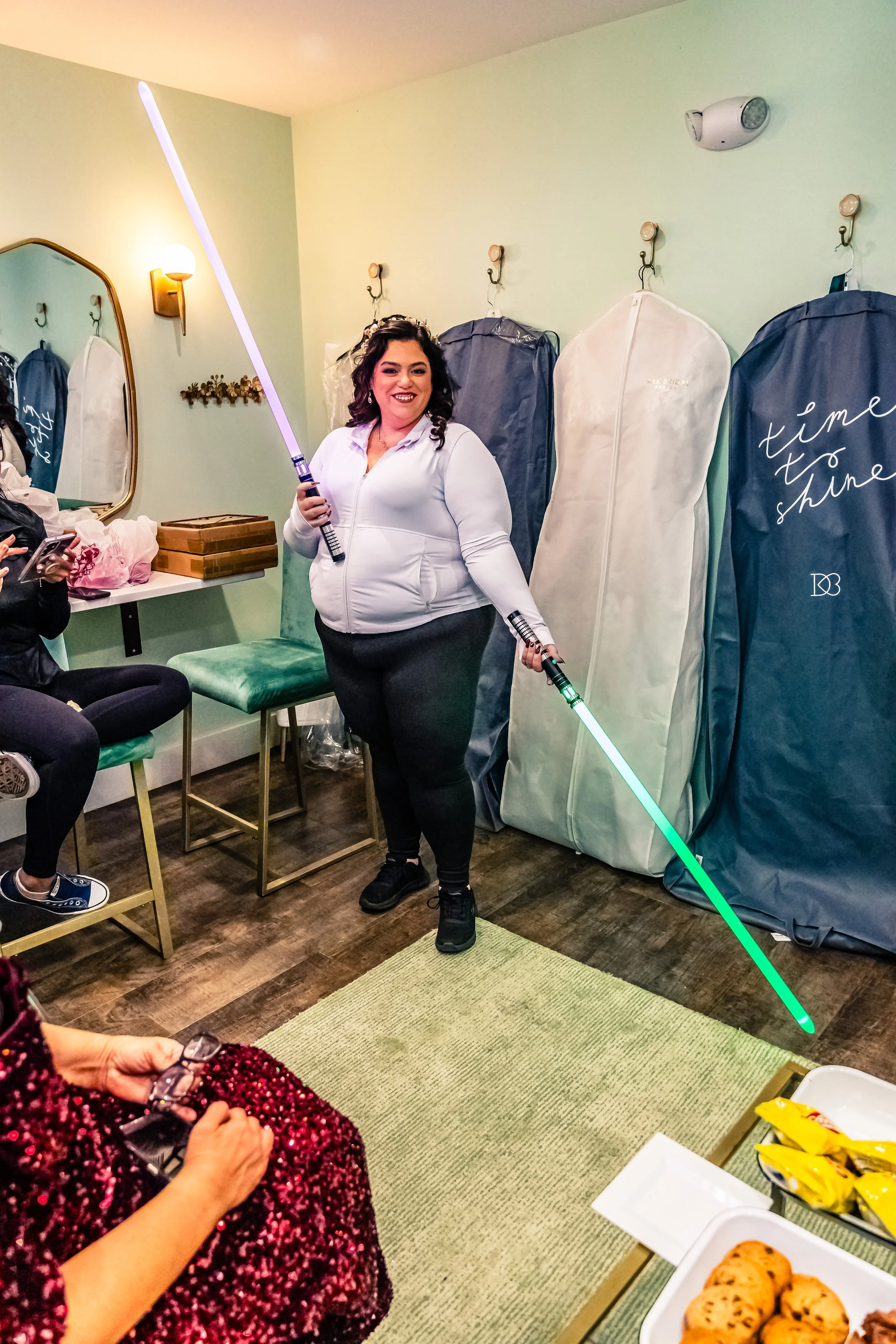 A woman is standing in a room, smiling, and holding two illuminated light sabers, one in each hand. She is wearing a white zip-up jacket and black pants. There are large items covered in protective covers against the wall behind her, and people are s