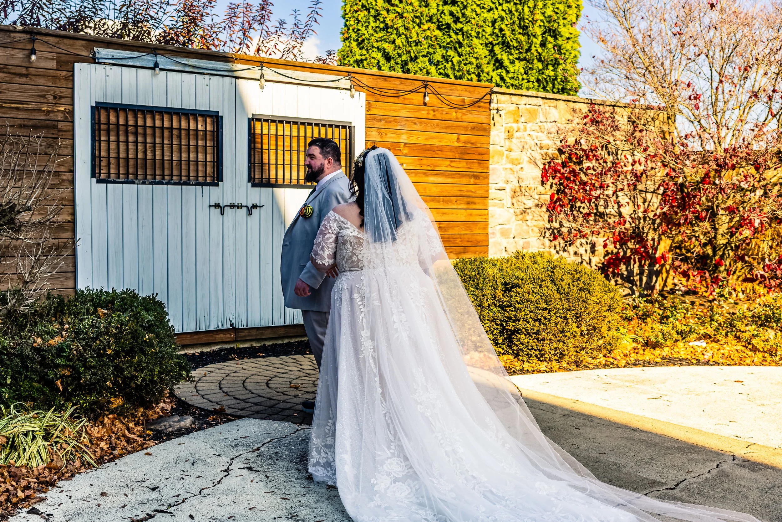 Bride and groom standing back-to-back outdoors in front of a wooden garage door, surrounded by autumn foliage, with the bride wearing a white lace wedding gown and veil, and the groom in a light gray suit.