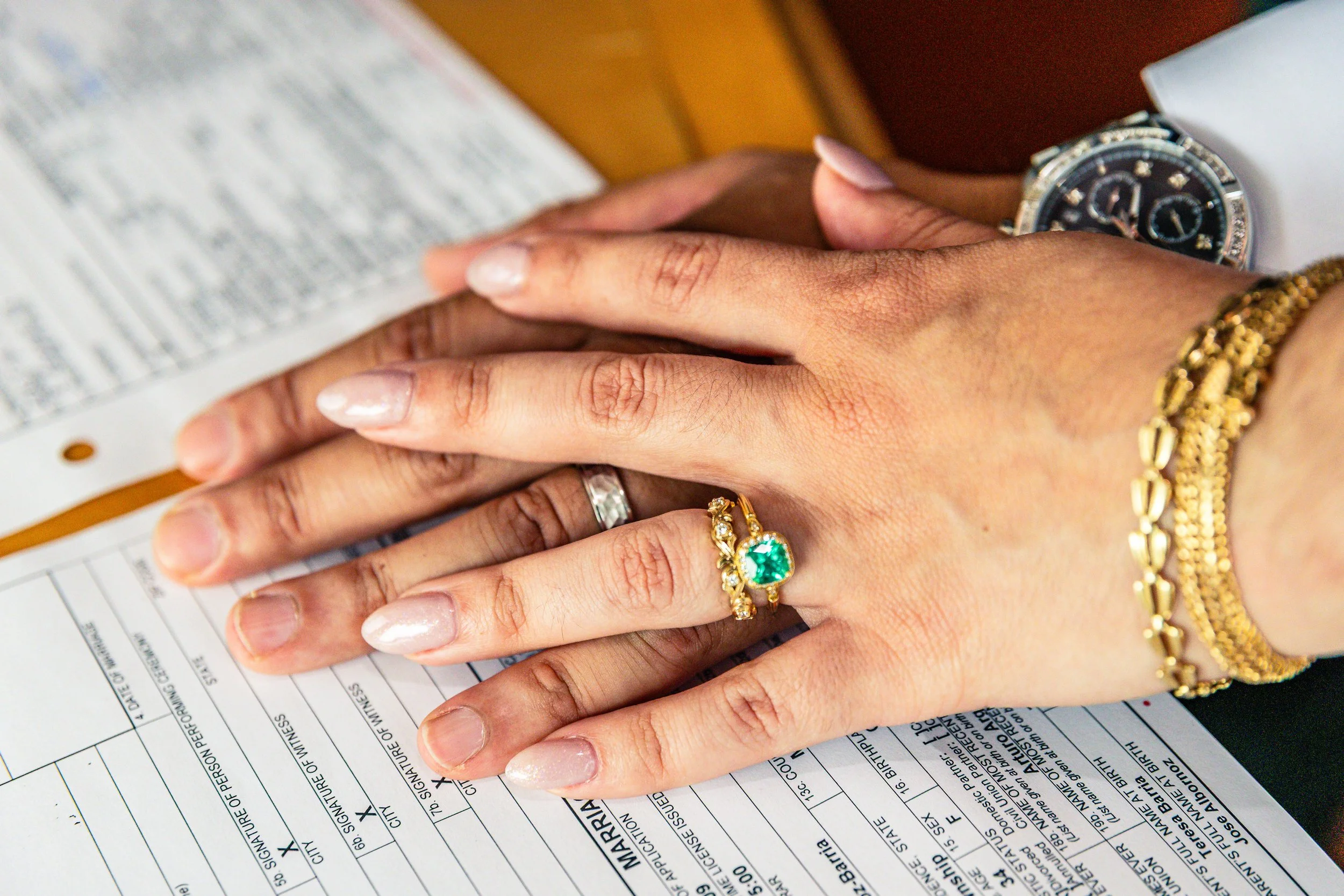 Two hands placed on a form or document, with the person wearing a watch, gold bracelets, and a ring with a green gemstone. The hand on top has a wedding band.
