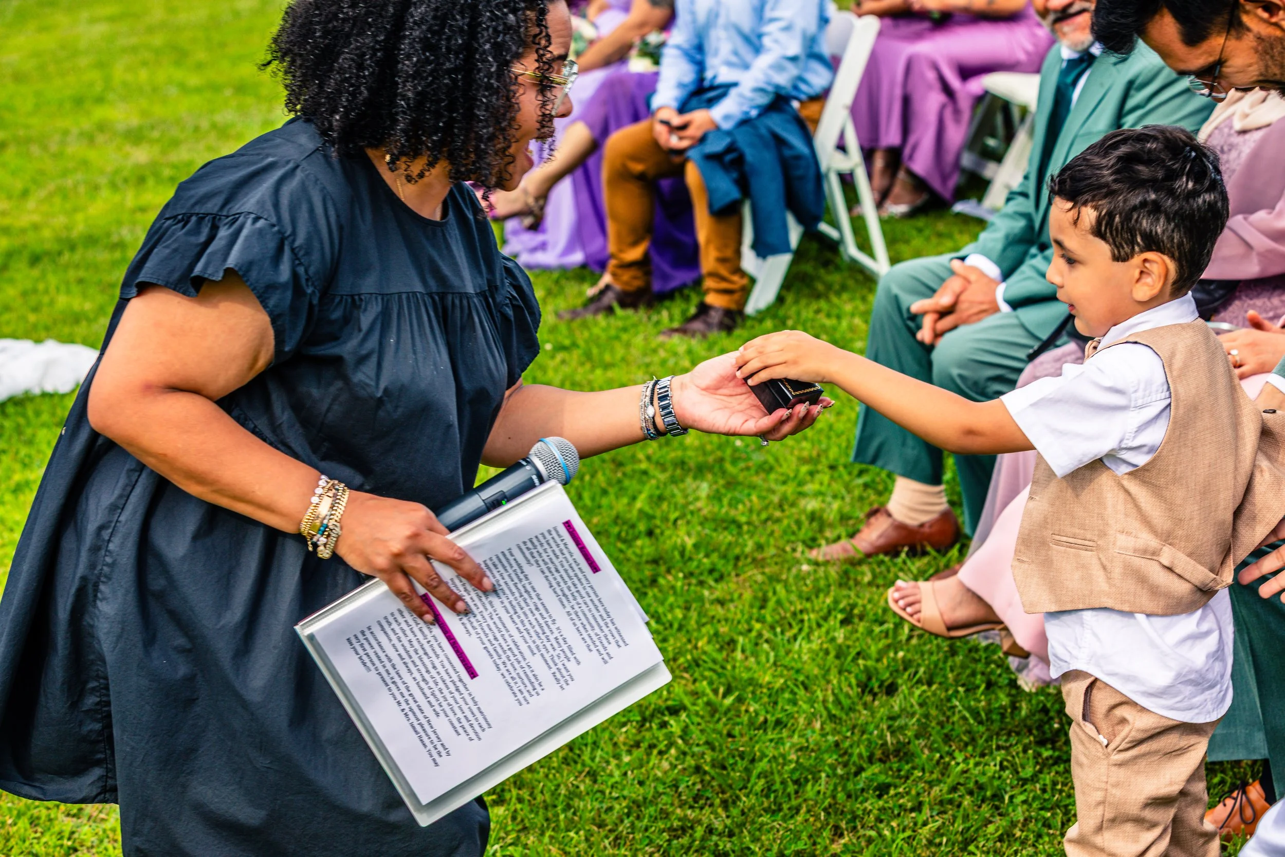 A woman in a black dress is handing a small box to a young boy at an outdoor event, with seated adults in the background.