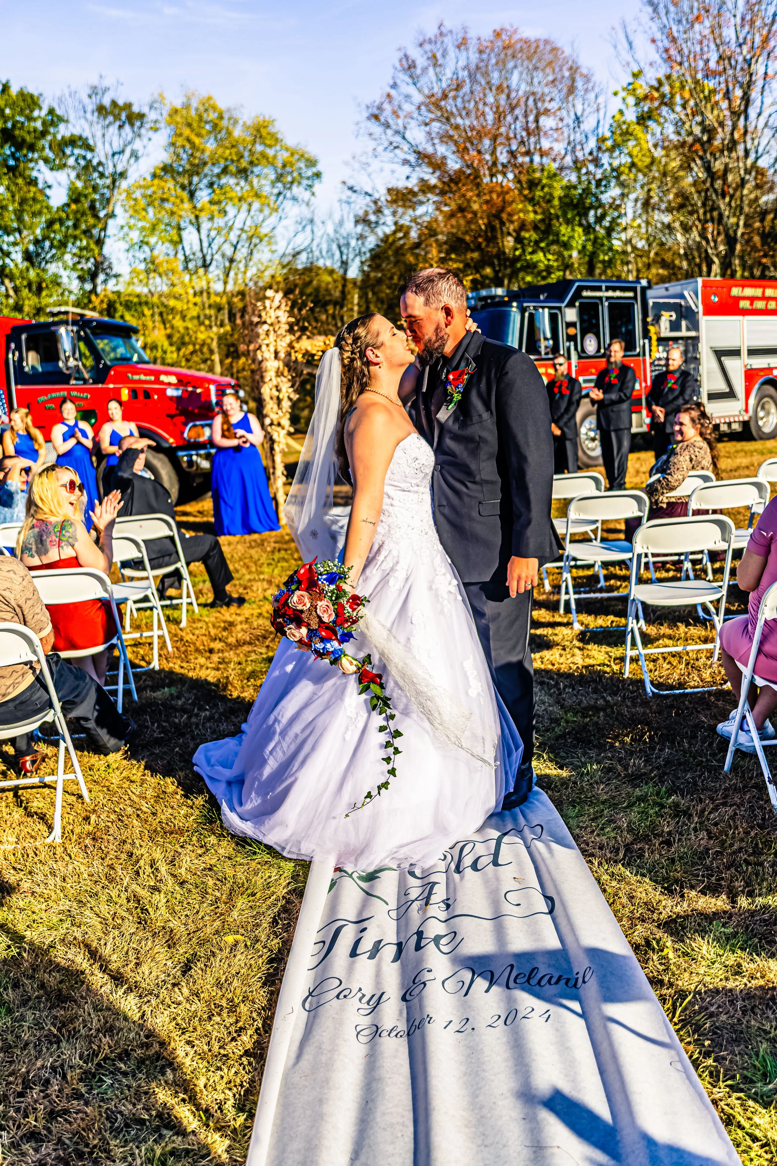 A bride and groom sharing a kiss during their outdoor wedding ceremony, with fire trucks and guests seated on white chairs in the background, on a sunny autumn day.