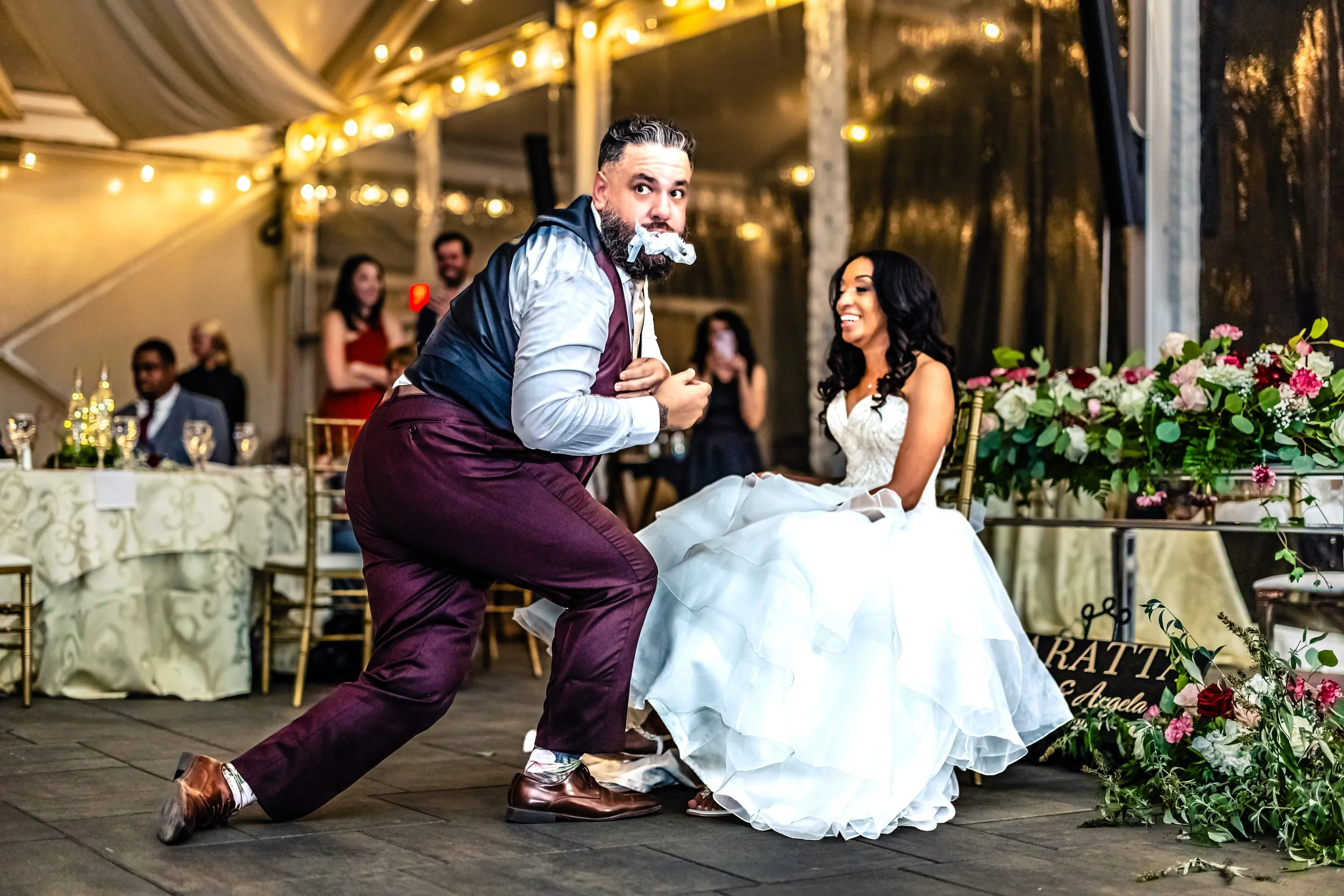 A man with a beard, wearing a gray shirt and maroon pants, with a napkin in his mouth, is dancing in front of a woman in a wedding dress, who is sitting at a decorated table with flowers. They are at a wedding reception under a tent with string light