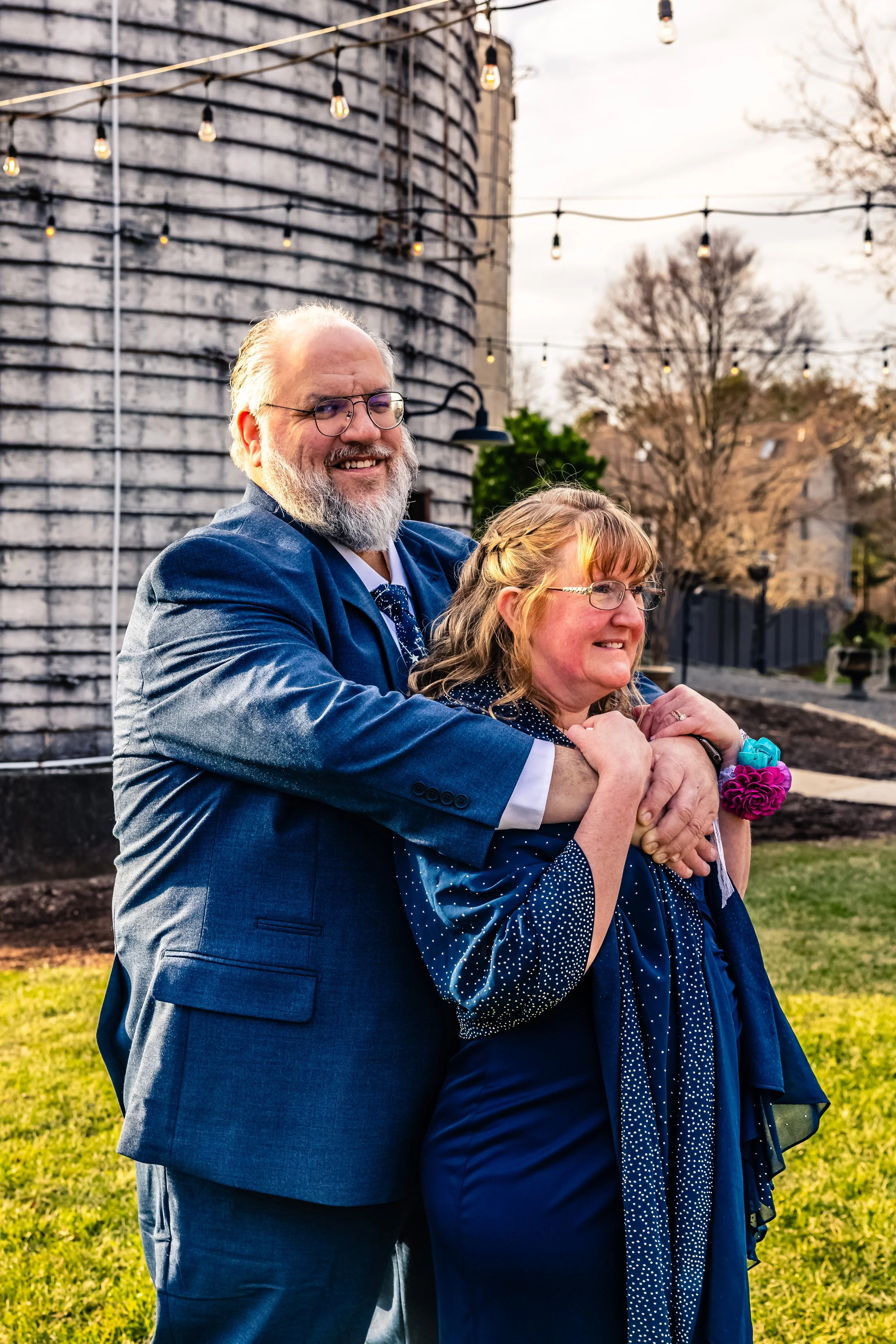 A happy man in a suit hugging a smiling woman in a dress outdoors during sunset, with string lights and a silo in the background.