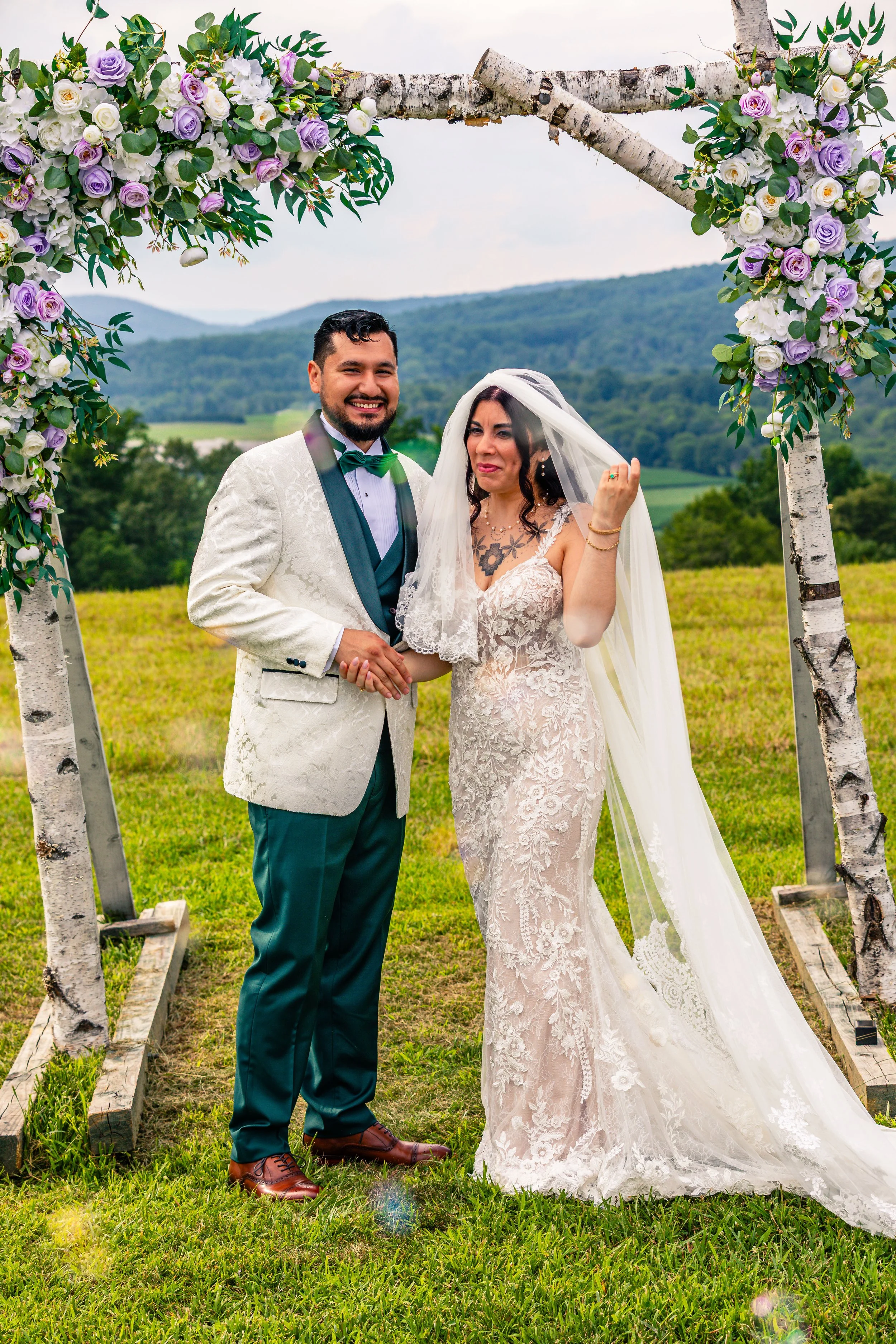 A newlywed couple on their wedding day standing beneath a decorated arch outdoors with green hills in the background. The groom is wearing a white suit jacket with a teal bow tie and teal pants, and the bride is in a lace wedding dress with a veil. T