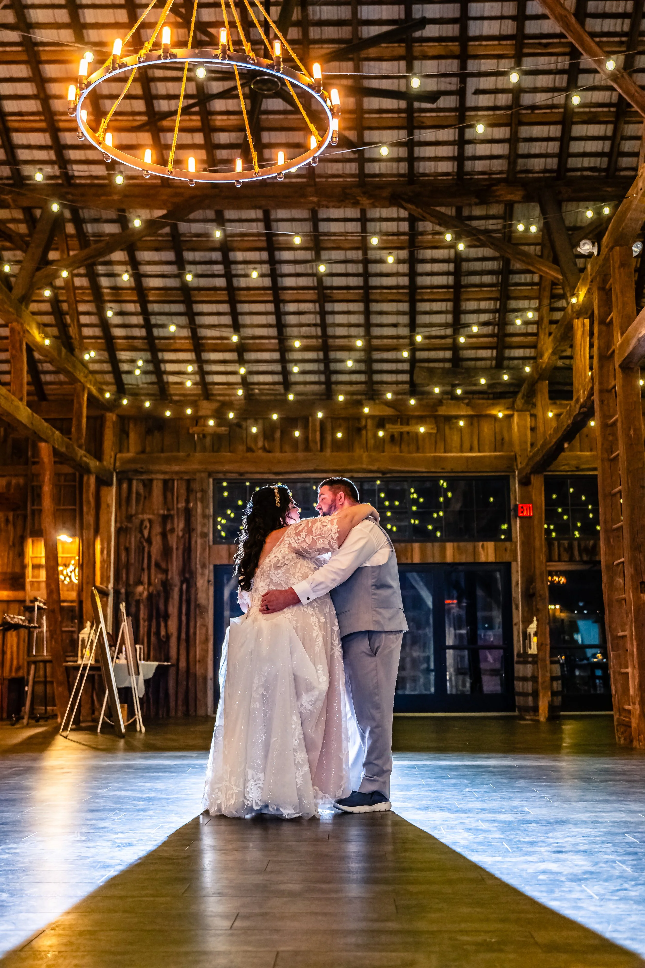 A bride and groom dancing in a rustic barn decorated with string lights and a large circular chandelier.