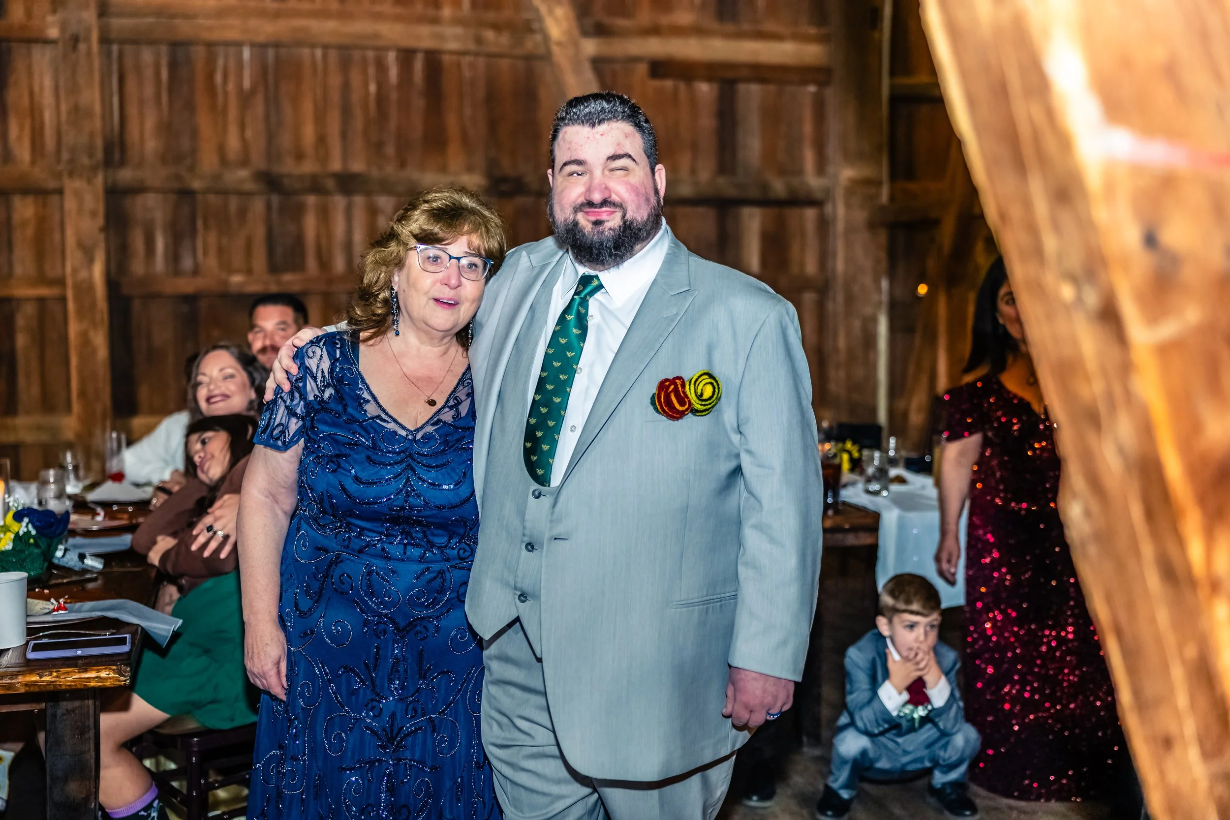 A man and a woman posing together at a social event in a rustic wooden venue, with several other people seated at tables in the background.