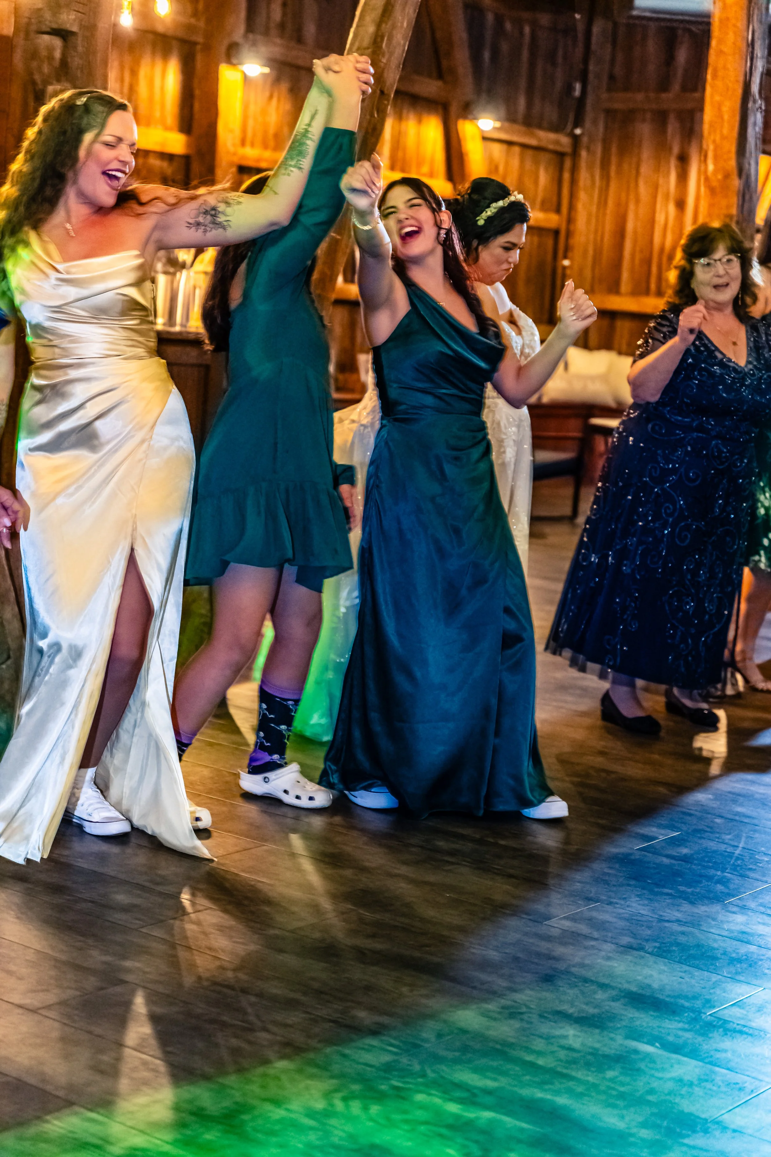 Group of women dancing and celebrating at a rustic indoor event, wearing elegant dresses and joyful expressions.
