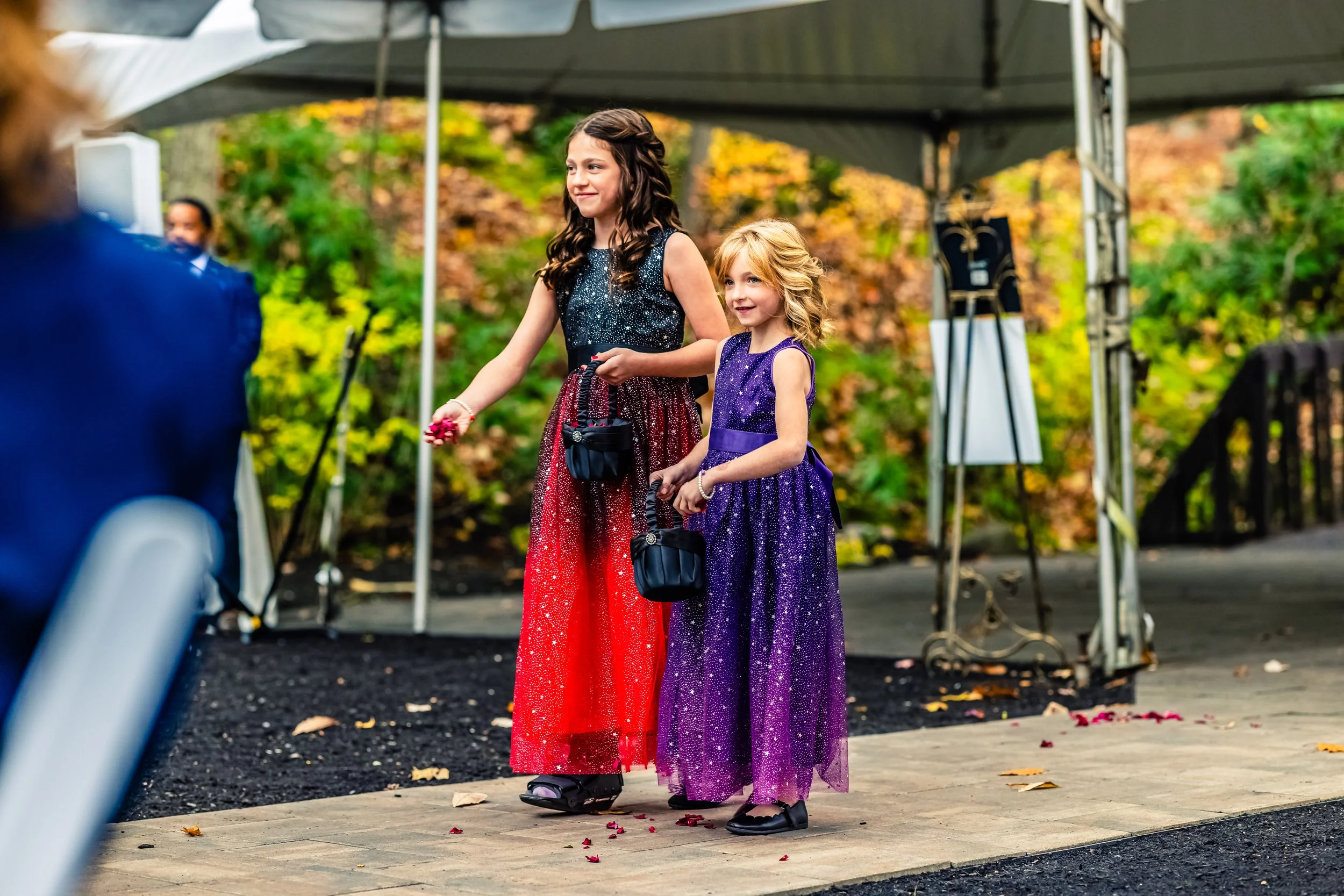 Two young girls in colorful, glittery dresses attending an outdoor formal event, holding small black bags, with a backdrop of trees with autumn leaves.