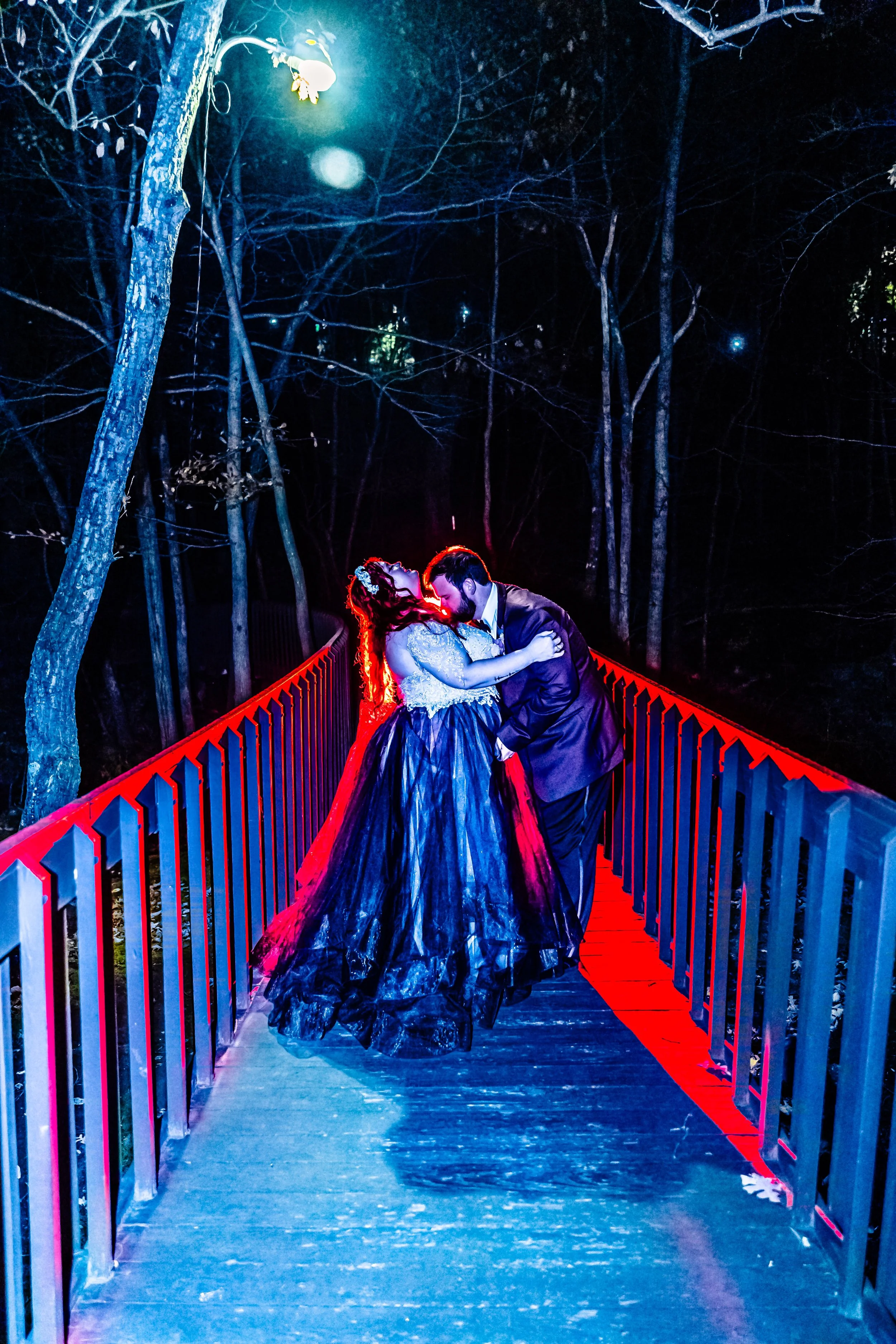 A couple dressed in formal attire sharing a kiss on a bridge at night, illuminated by colorful neon and street lighting, with trees in the background.