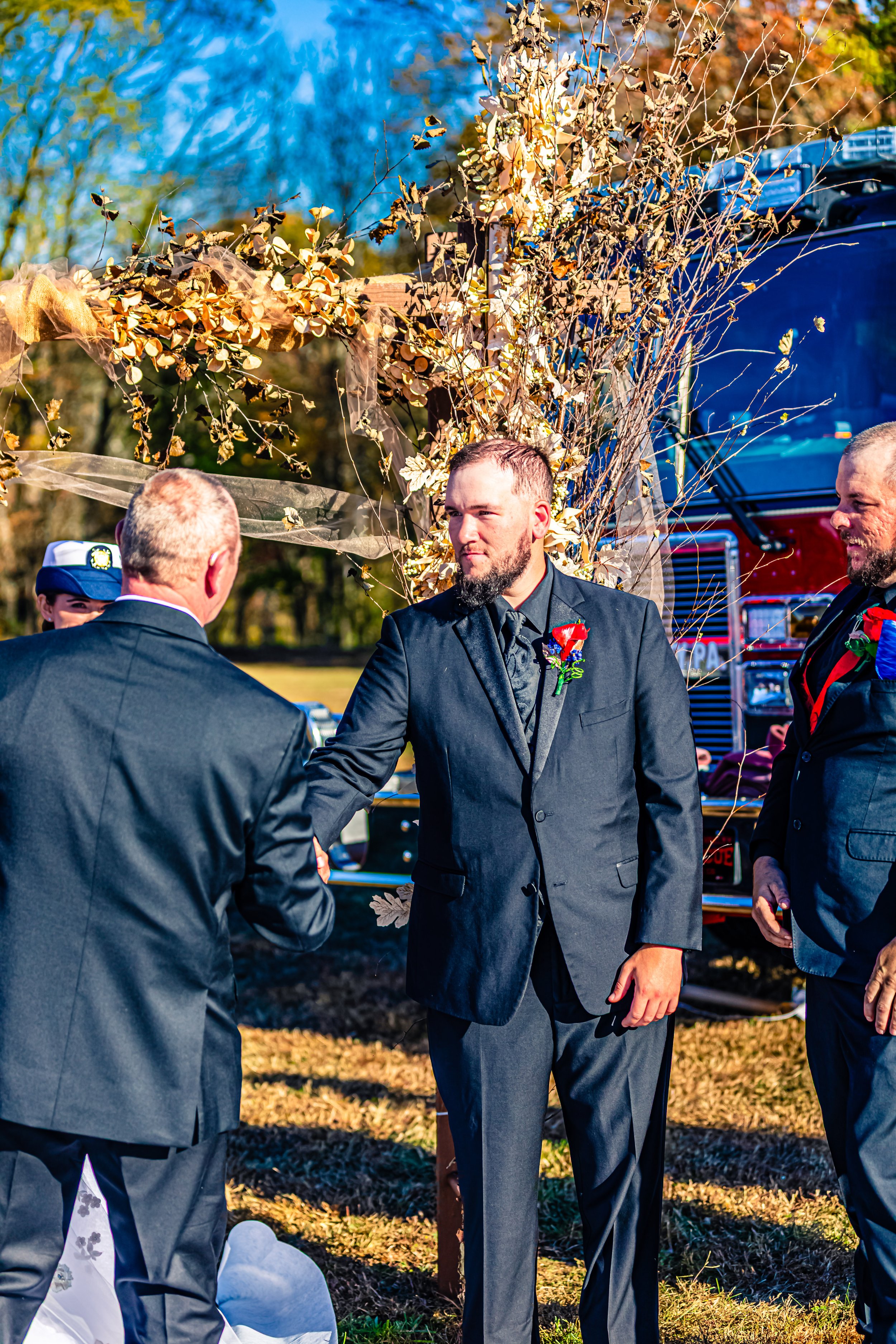Man in a black suit shaking hands with an older man during a wedding ceremony outdoors, decorated with dried foliage and flowers, with a fire truck in the background.