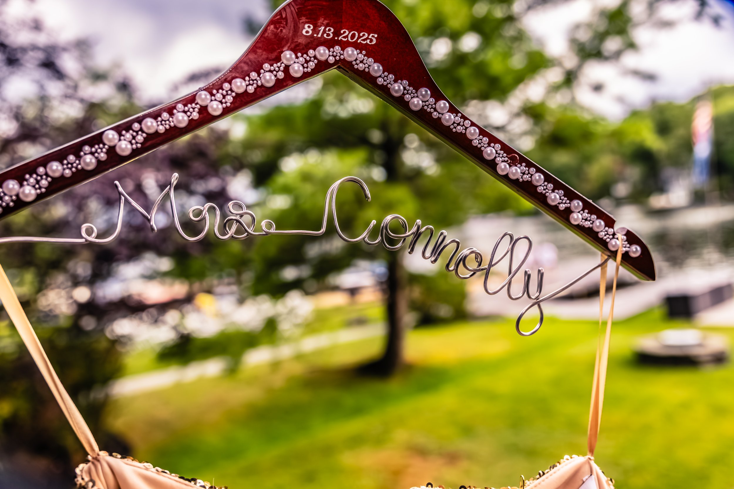 A decorative hanger with the words 'Mrs. Cannonly' crafted in wire, decorated with small pearl beads, and a date '8.13.2025' written on the top part of the hanger, set outdoors with blurred greenery in the background.