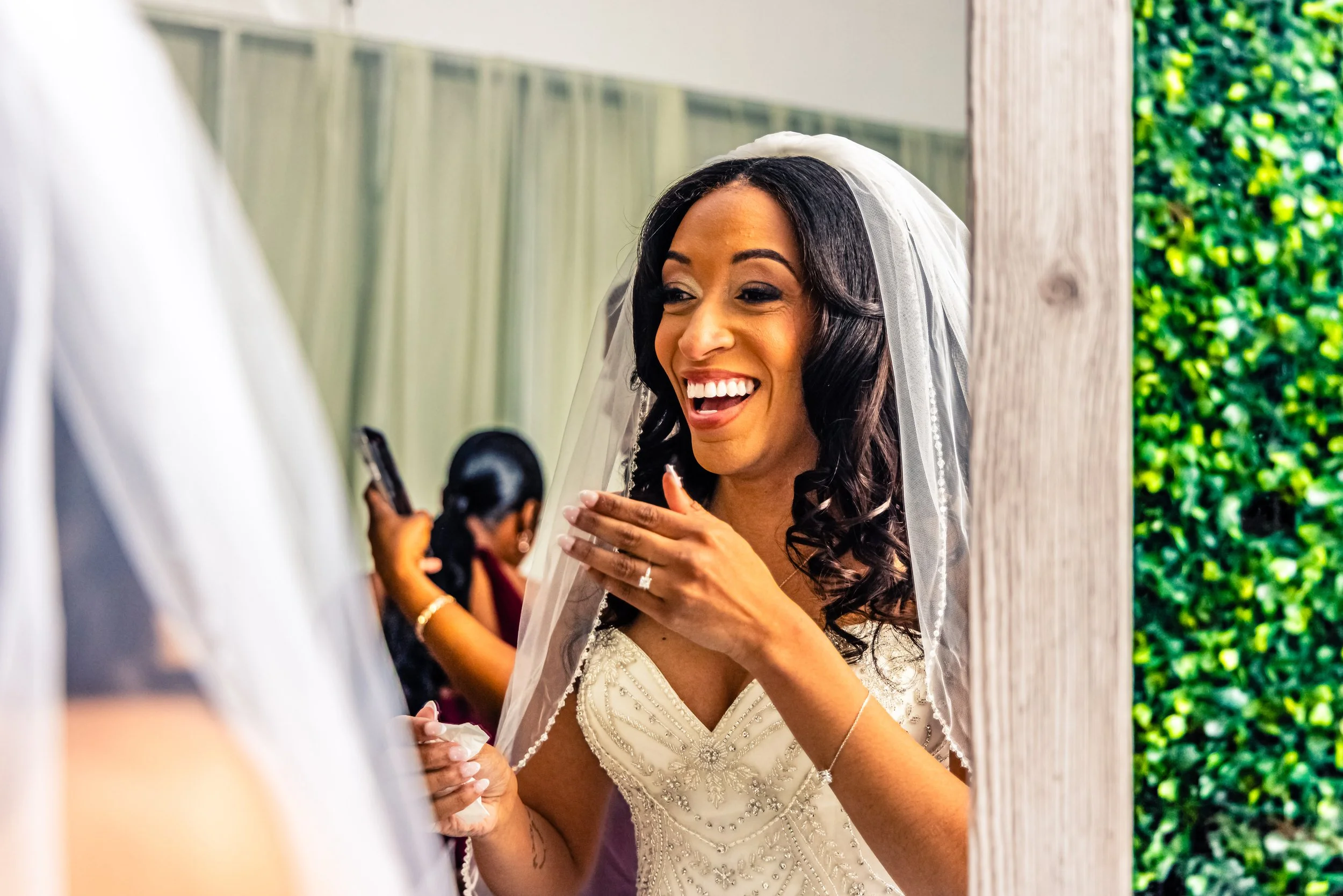 A bride with dark hair in a white wedding dress and veil, smiling and touching her face, looking into a mirror.