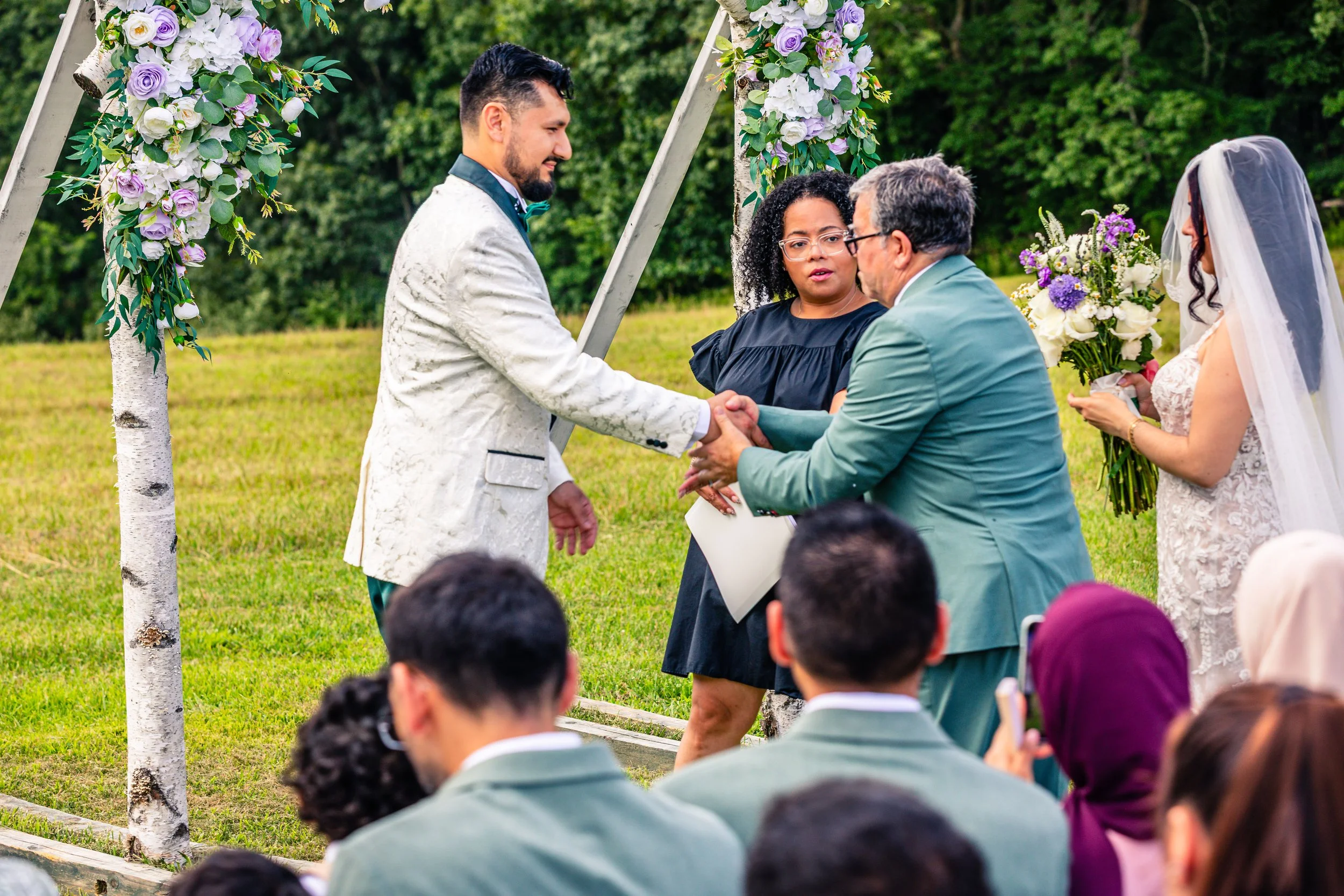 A wedding ceremony outdoors with a couple exchanging vows, a officiant, and a bride holding a bouquet, under a floral arch with guests seated nearby.