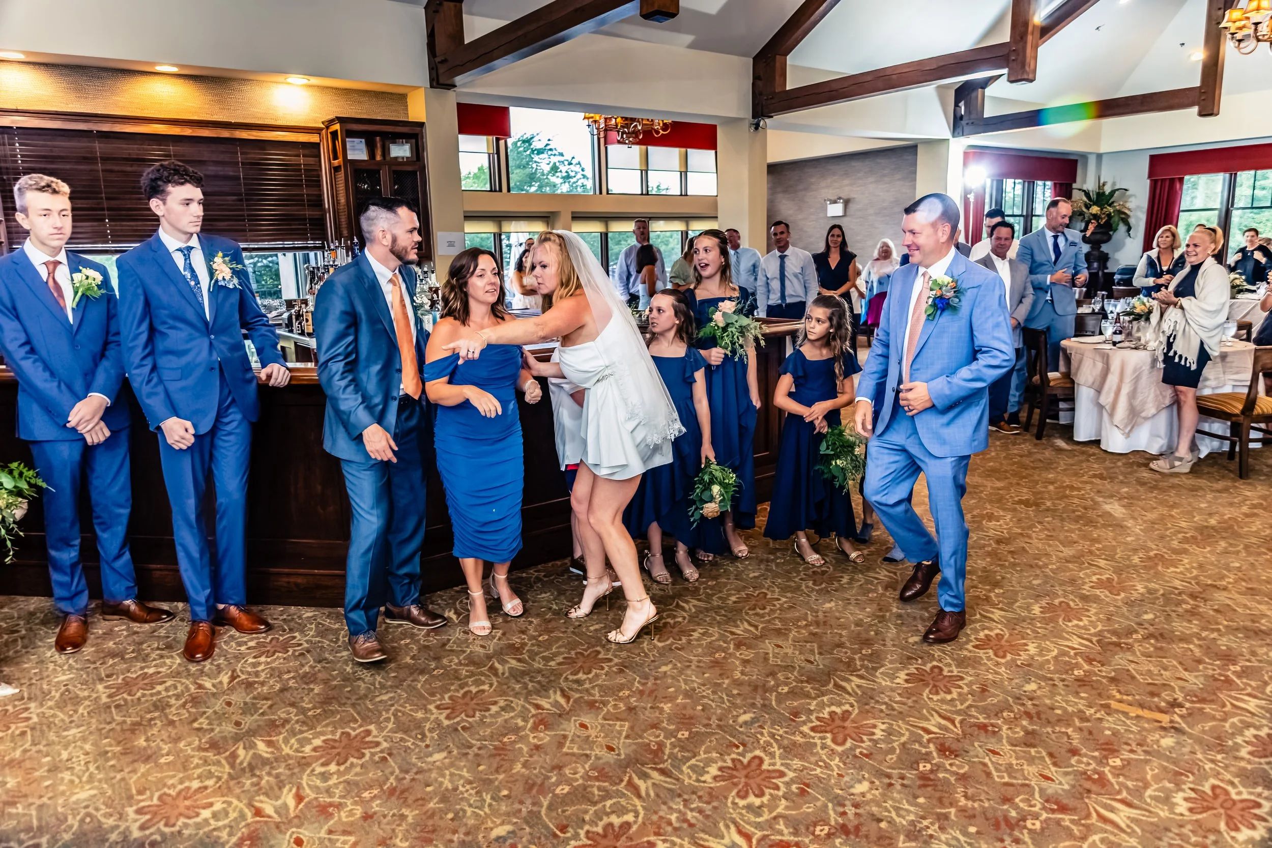 Group of wedding guests dressed in blue suits and dresses, gathered indoors at a wedding reception. Some people are dancing and chatting near the bar area, with tables and windows in the background.