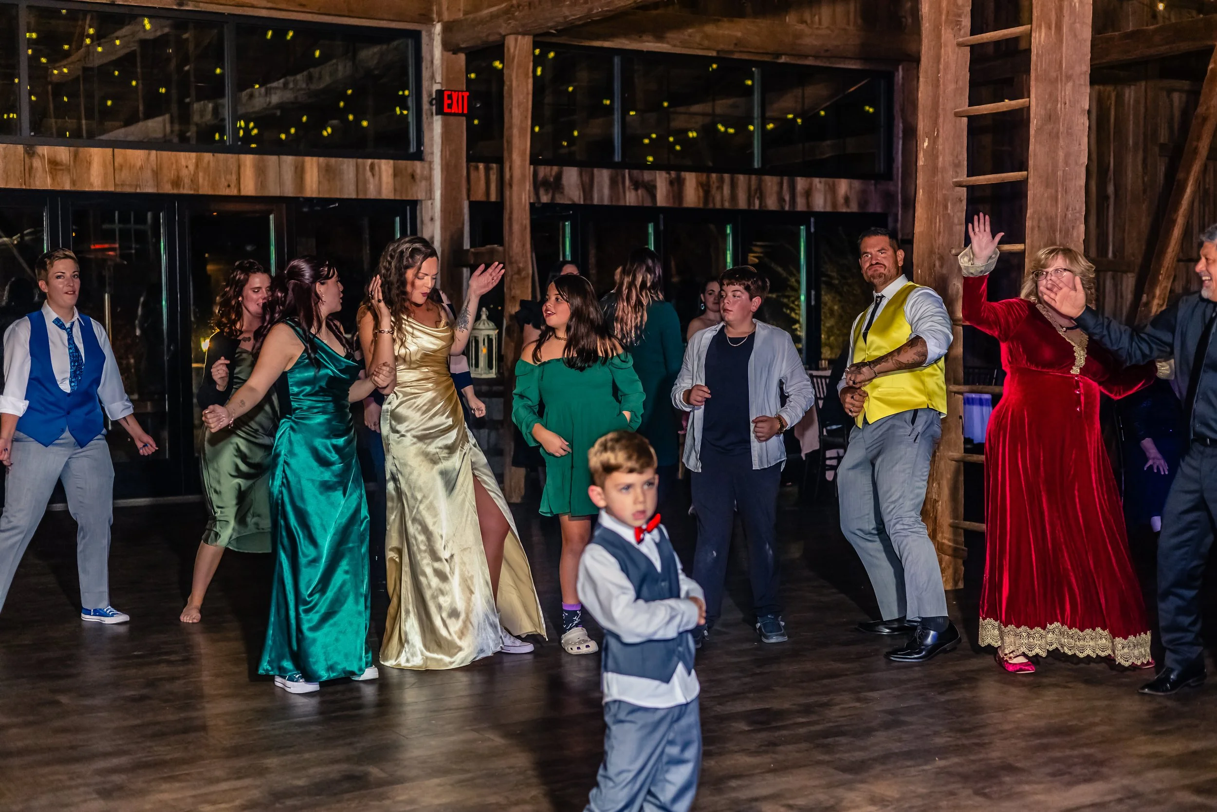 People dancing at a party or celebration in a rustic venue with wooden beams and walls, some dressed in formal attire, with a young boy in the foreground wearing a vest and bow tie.