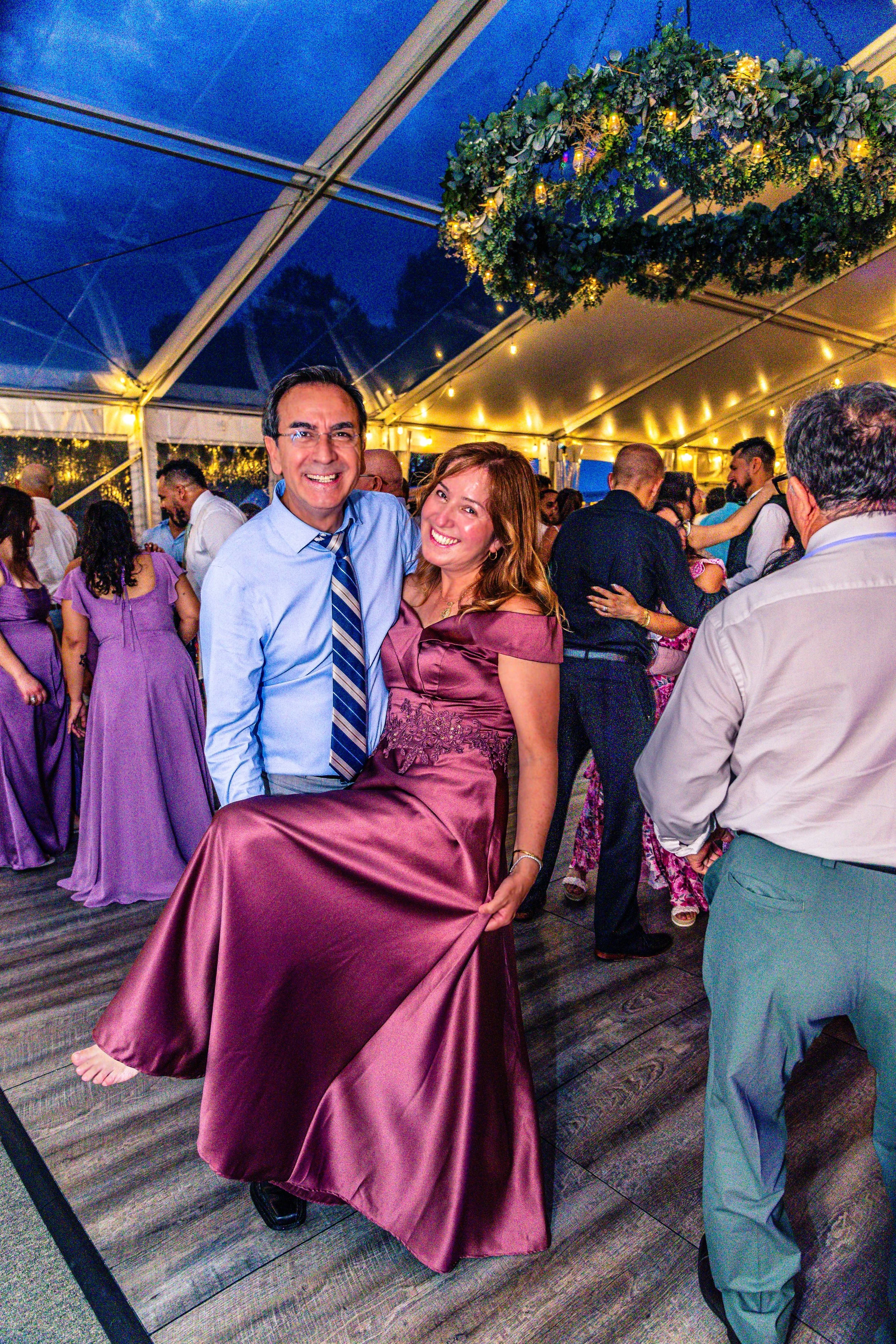 A man and woman dancing at a lively celebration or party under a tent decorated with string lights and a floral arrangement hanging from the ceiling, with other guests dancing in the background.