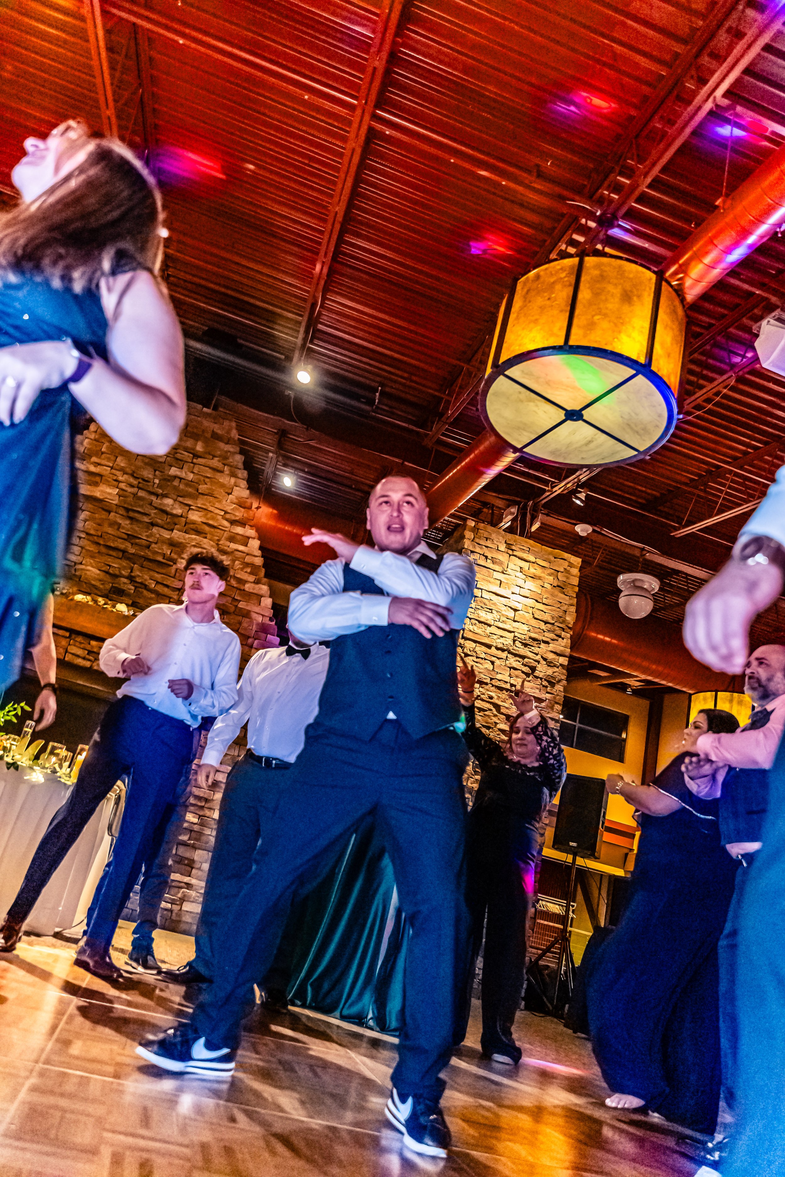 People dancing at a party in a restaurant or ballroom with brick walls and colorful lighting.