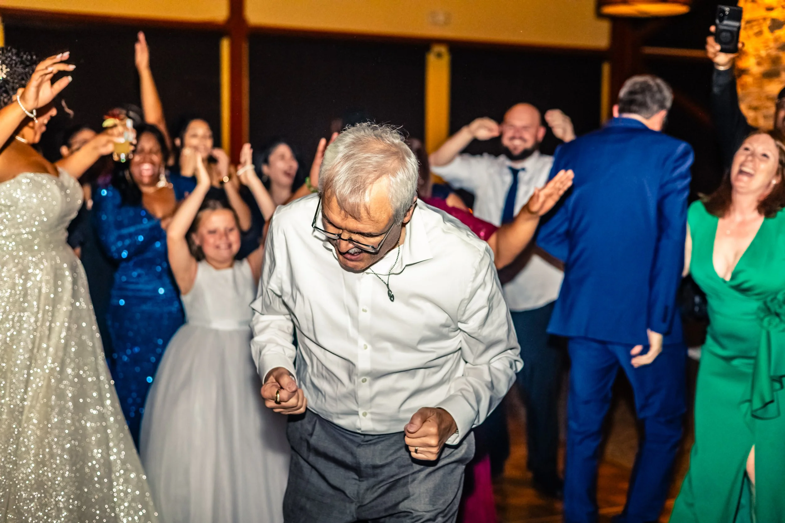People dancing and celebrating at a festive party or wedding reception, with a man in the foreground cheering and others in colorful attire enjoying the moment.