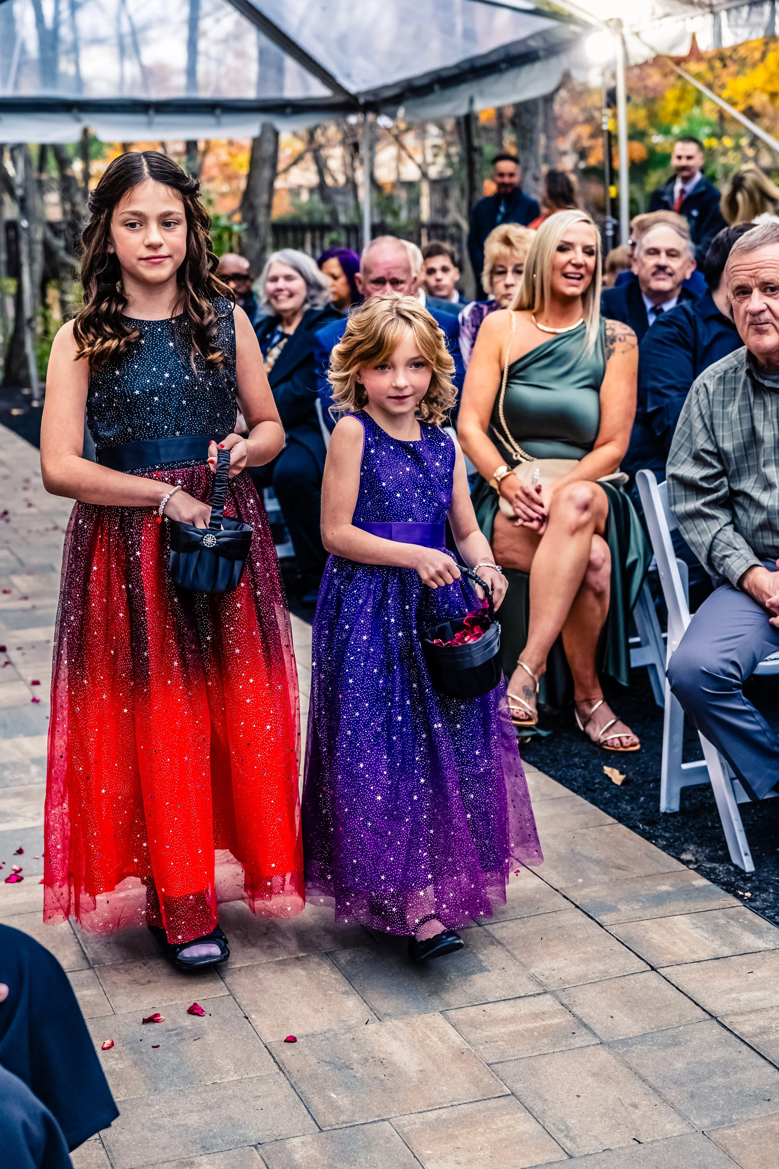 Two young girls in colorful, sparkly dresses walking down the aisle at an outdoor wedding ceremony, surrounded by seated guests.